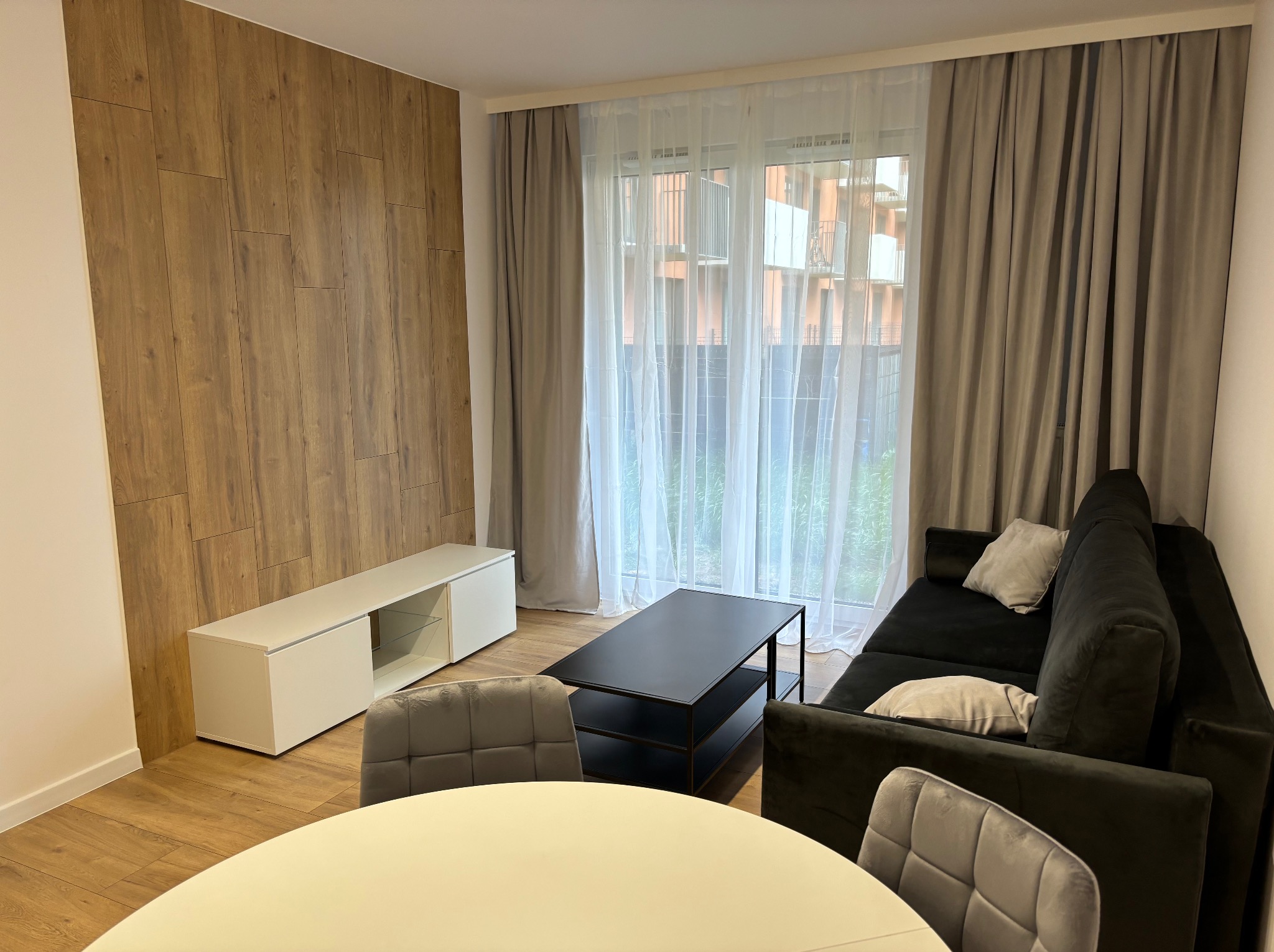 Nowoczesny salon z drewnianą ścianą, szarą sofą i białymi meblami. Widok na okno z zasłonami. Stół z krzesłami na pierwszym planie.