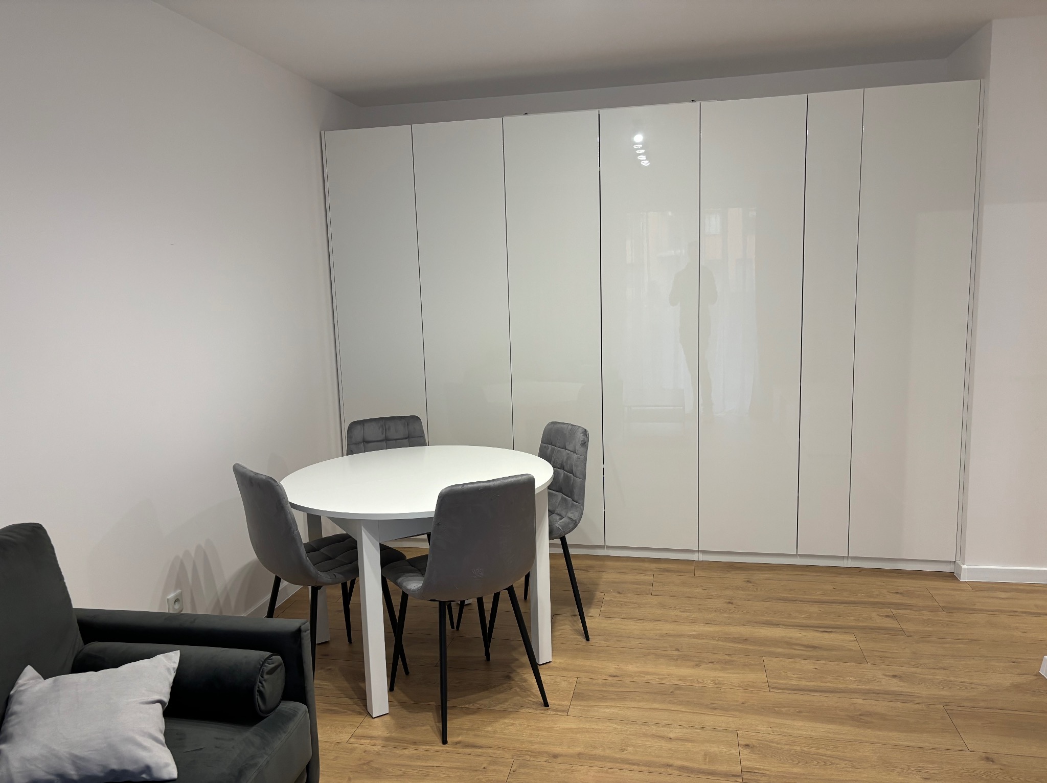 Jasne wnętrze: biały stół z szarymi krzesłami na panelach, na tle ściany z białymi, błyszczącymi szafami w zabudowie. W rogu widoczny fragment szarej sofy.