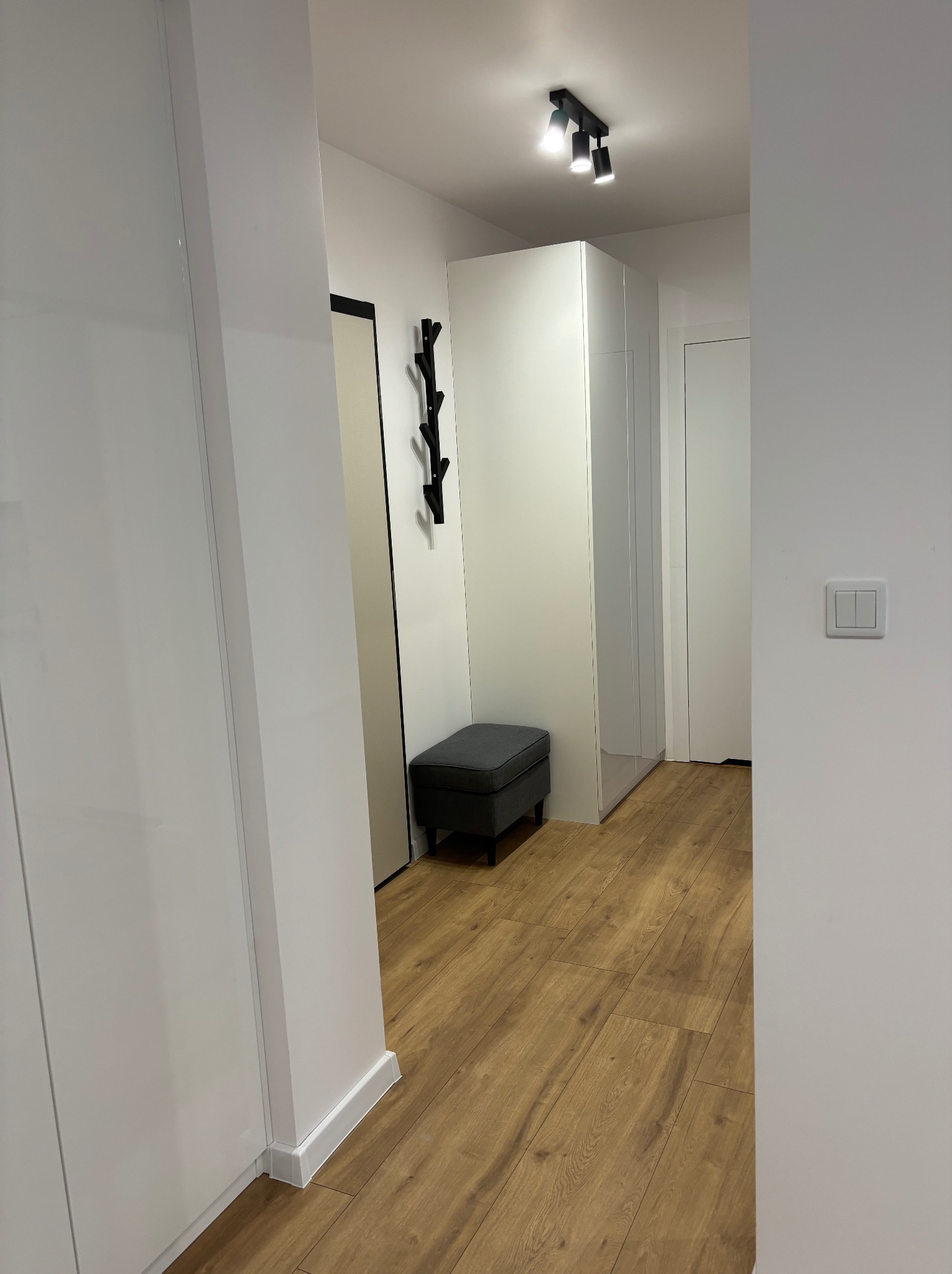 Nowoczesny przedpokój z białymi szafami, drewnianą podłogą i szarą pufą. Minimalistyczny wieszak na ścianie. Oświetlenie sufitowe punktowe.
