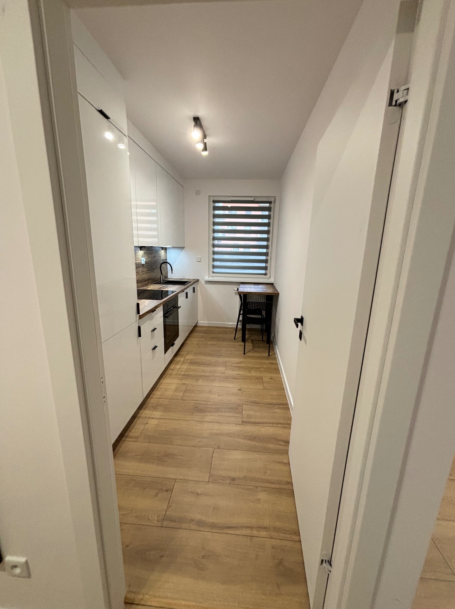 Wnętrze wąskiej, nowocześnie urządzonej kuchni z białymi szafkami, drewnianym blatem i podłogą, widzianej z perspektywy wejścia. W tle okno z żaluzjami.