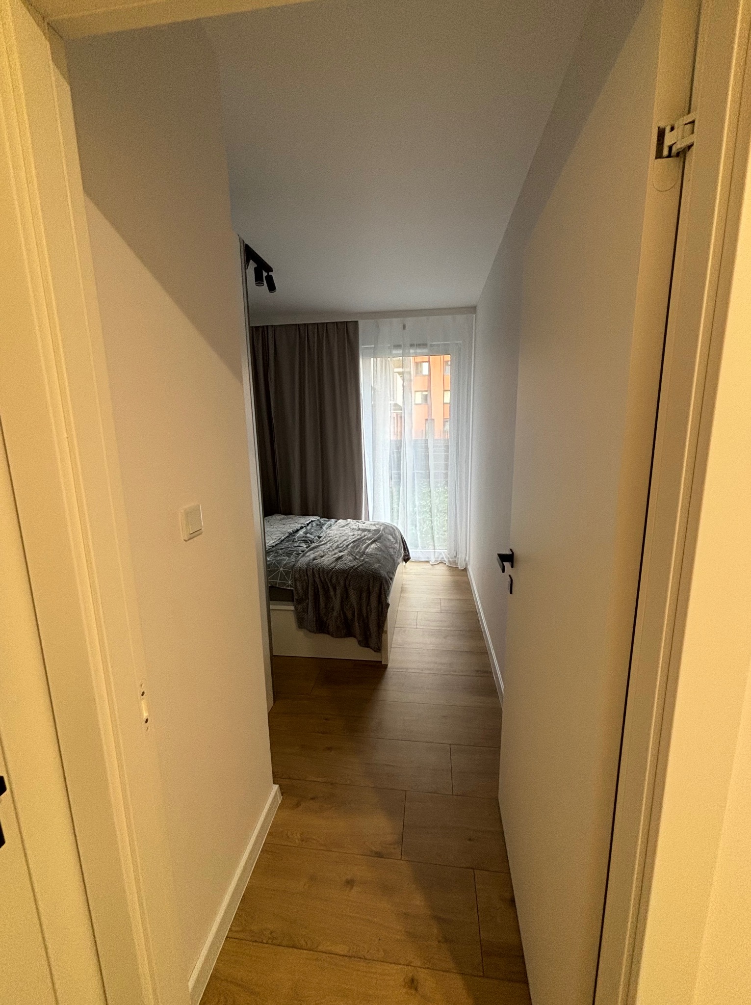 Widok z korytarza na sypialnię z drewnianą podłogą i łóżkiem z szarą narzutą. Białe ściany i drzwi. Minimalistyczne oświetlenie sufitowe.