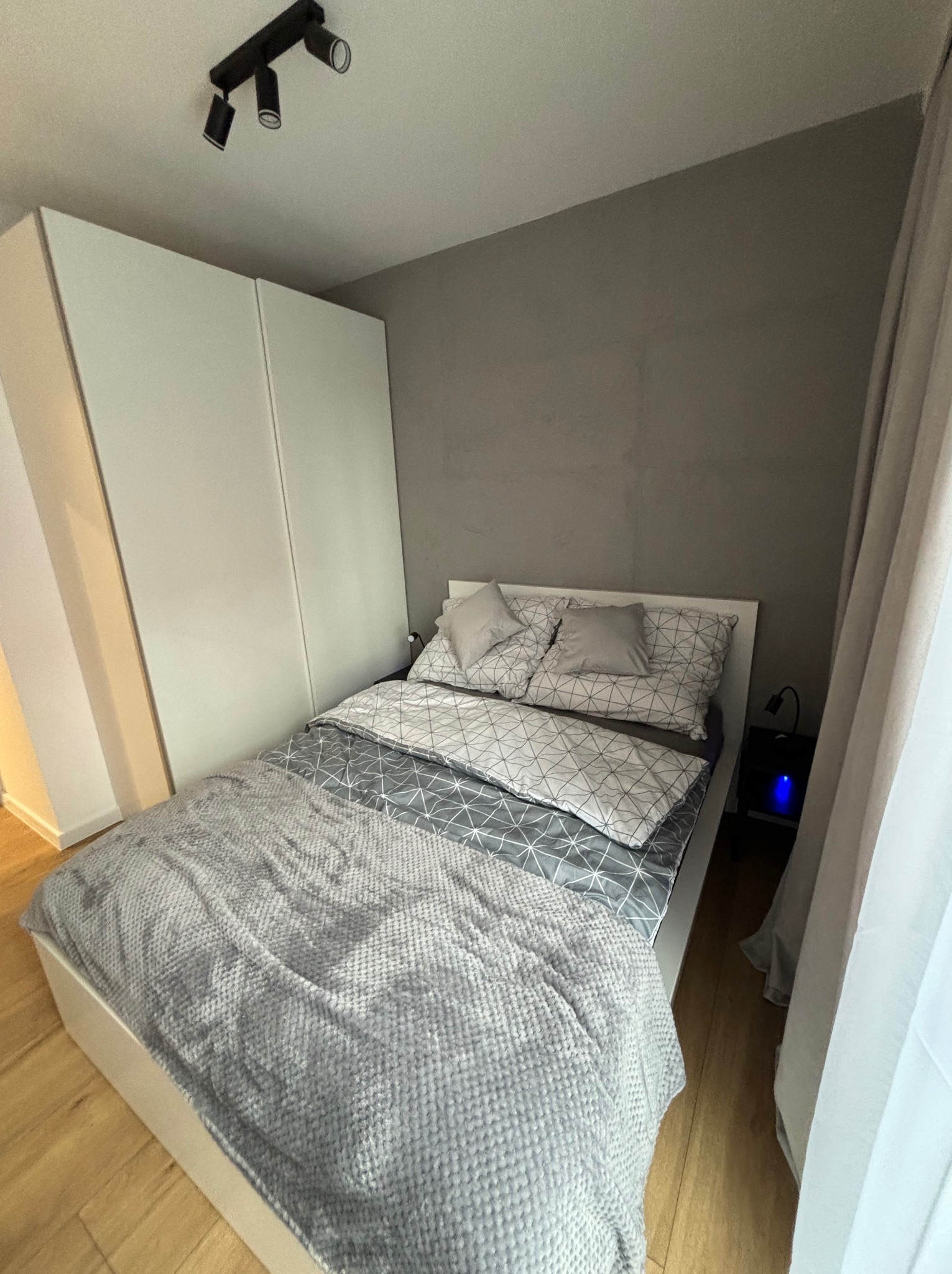 Nowoczesna sypialnia z białym łóżkiem, szarą pościelą w geometryczne wzory i dużą białą szafą. Minimalistyczne oświetlenie sufitowe i drewniana podłoga dodają przytulności.