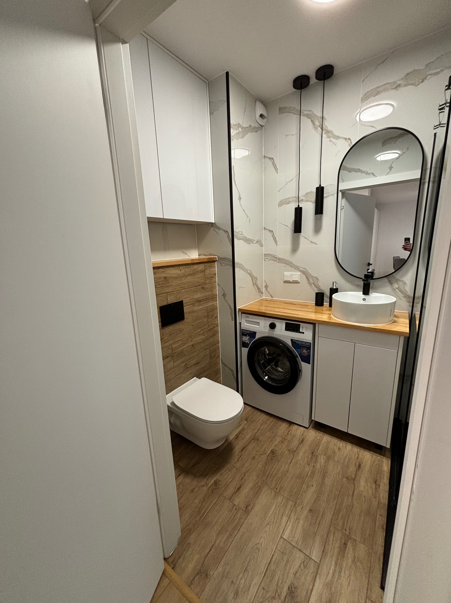 Nowoczesna łazienka z marmurowymi płytkami, drewnianym blatem, owalnym lustrem i pralką pod umywalką. Minimalistyczne oświetlenie i podłoga imitująca drewno.