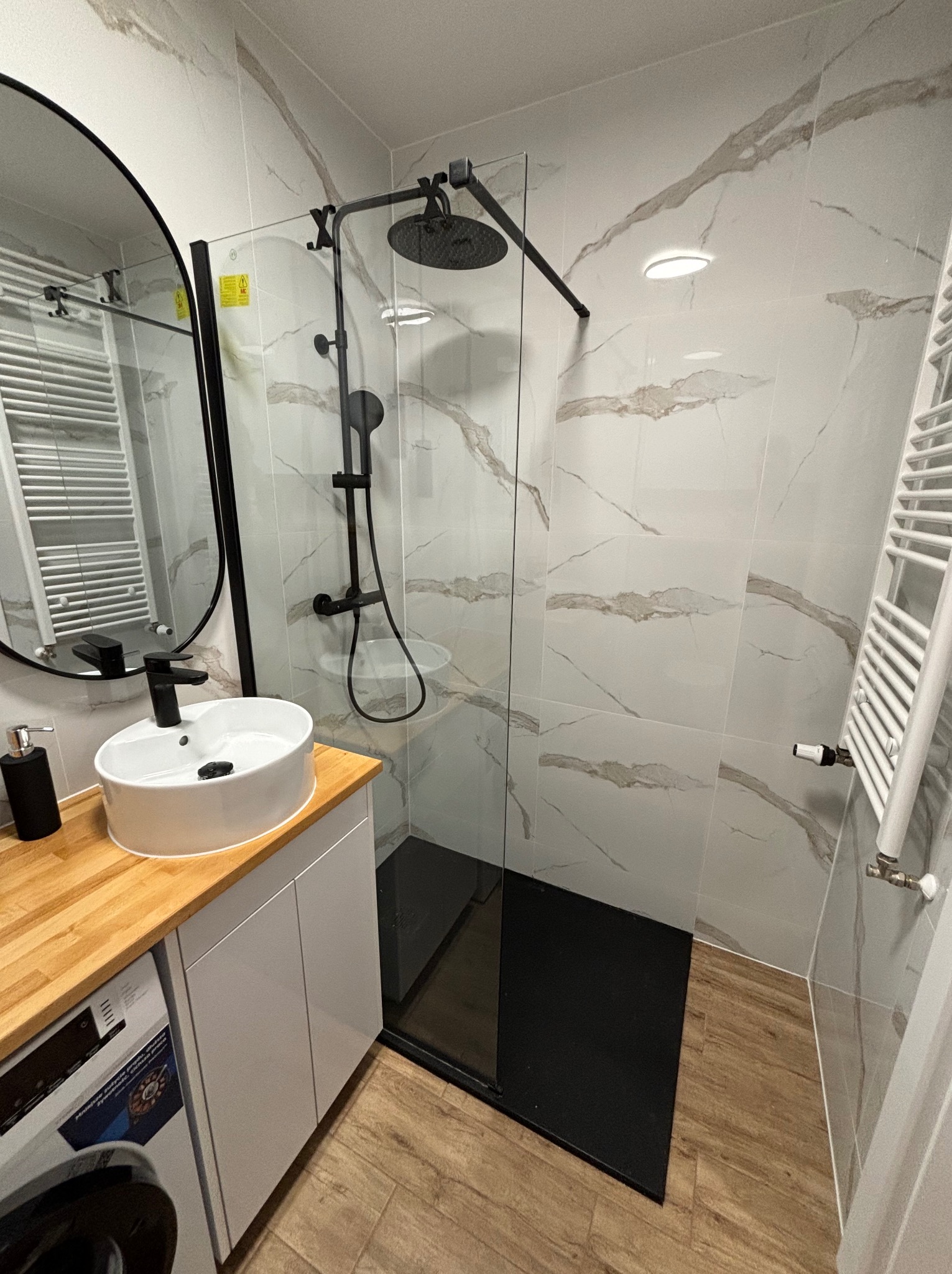Nowoczesna łazienka z kabiną typu walk-in, białą umywalką na drewnianym blacie i grzejnikiem drabinkowym. Marmurowe płytki na ścianach i czarny brodzik.