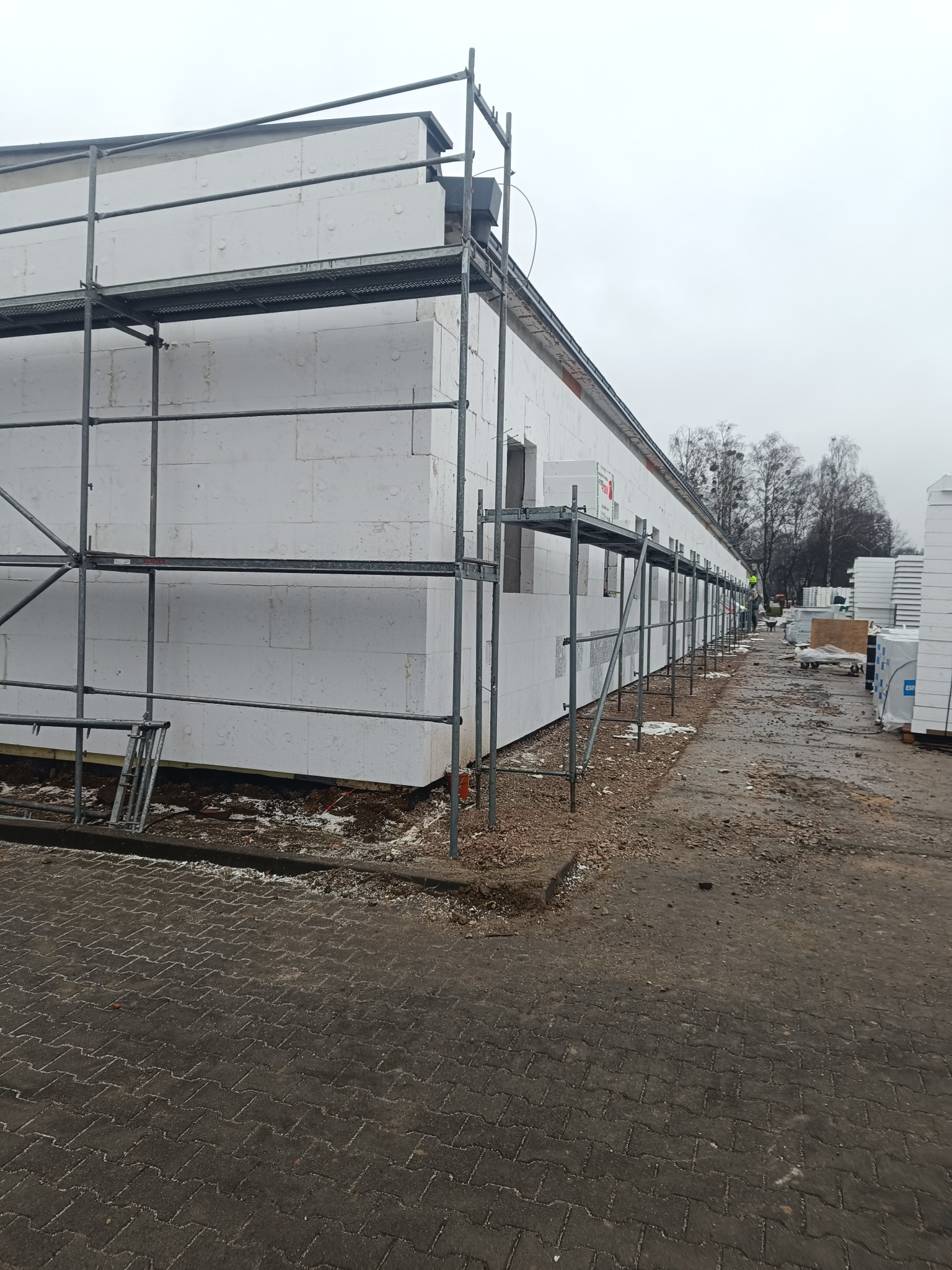 Budynek w trakcie ocieplania styropianem, widoczne rusztowania i materiały budowlane. Prace elewacyjne w toku, pochmurne niebo w tle.