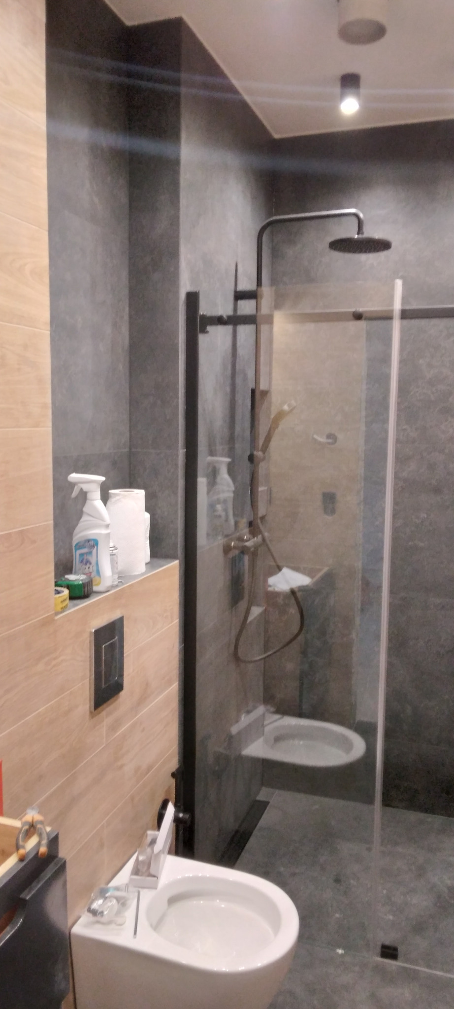 Nowoczesna łazienka po remoncie: szara glazura, kabina walk-in z czarnymi profilami, podwieszana toaleta i bidet. Na półce środki czystości.