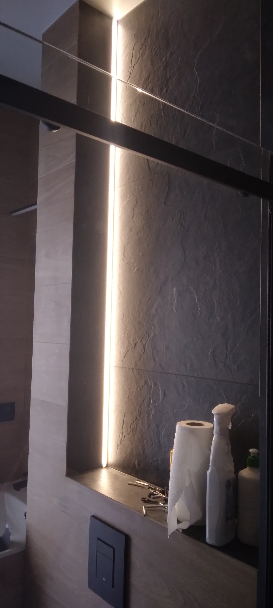 Nowoczesna łazienka z podświetleniem LED w pionowej wnęce. Ciemna, kamienna ściana kontrastuje z drewnopodobnymi płytkami. Na półce akcesoria łazienkowe i drobne narzędzia.