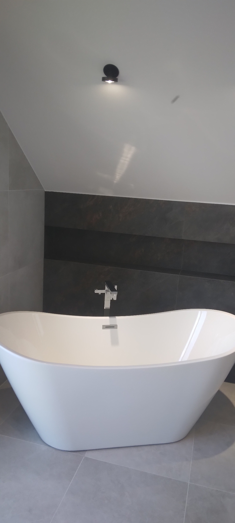 Nowoczesna, wolnostojąca wanna z chromowaną baterią na tle ciemnoszarych płytek i jasnego sufitu z punktowym oświetleniem. Minimalistyczny design łazienki.