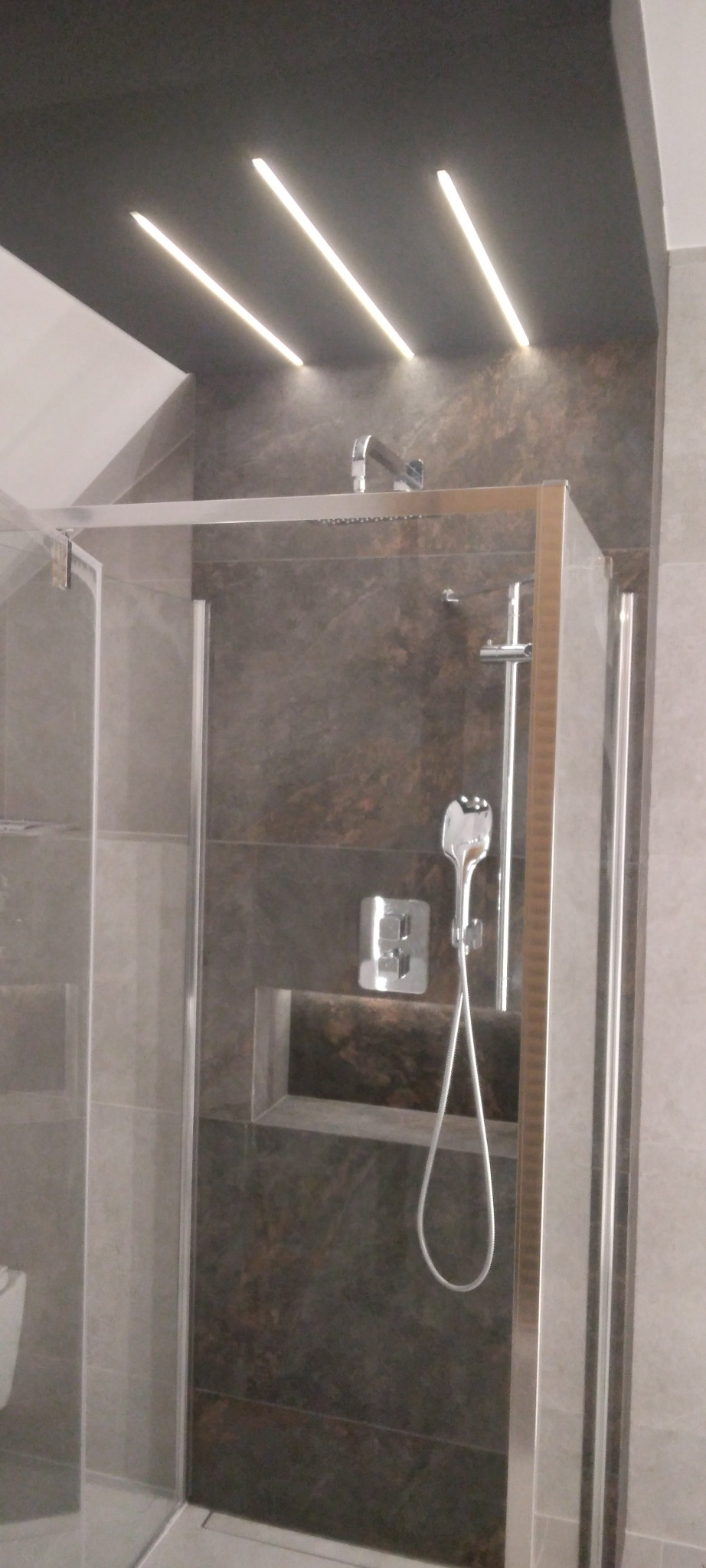 Nowoczesna kabina prysznicowa z deszczownicą i wnęką na kosmetyki, wykończona grafitową płytką imitującą kamień, oświetlona liniowymi lampami LED w suficie.