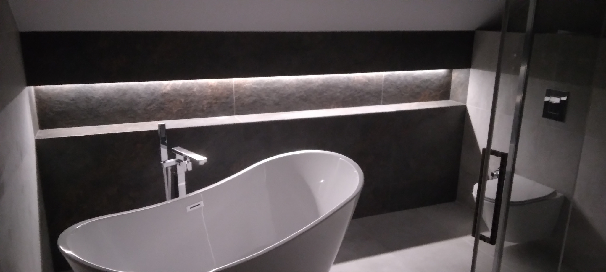 Nowoczesna łazienka z wolnostojącą białą wanną na tle ciemnej, podświetlonej wnęki. Minimalistyczny design z elementami szarości i bieli.