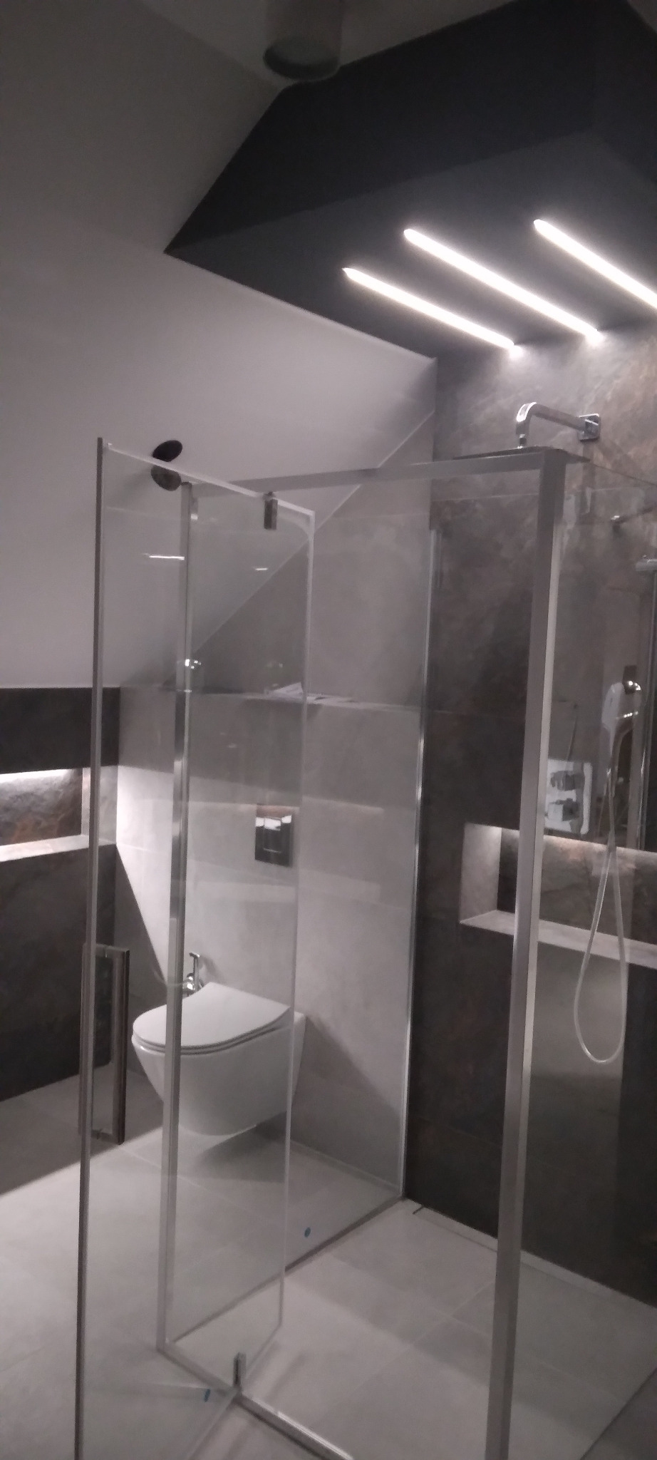 Nowoczesna łazienka z kabiną prysznicową typu walk-in, podświetlanym sufitem i toaletą w tle. Minimalistyczny design z szarymi płytkami i chromowanymi elementami.