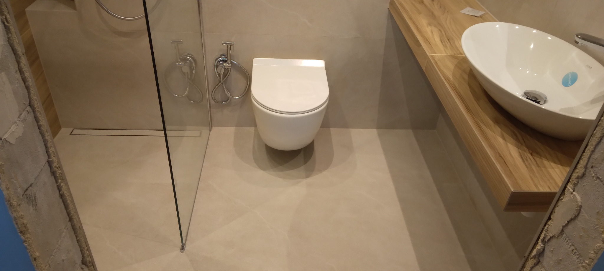 Nowoczesna łazienka z podwieszaną toaletą, bidetką i umywalką nablatową na drewnianym blacie. Widoczna szklana kabina prysznicowa i beżowe płytki.