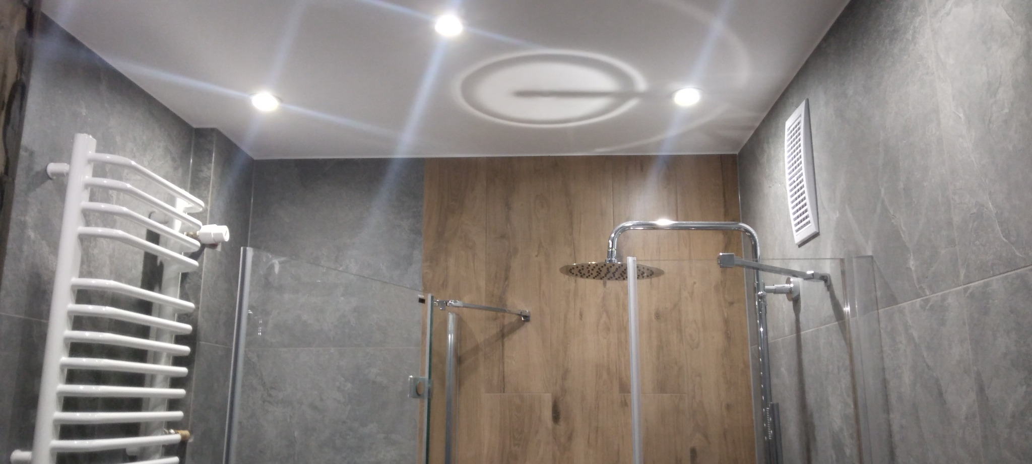 Nowoczesna łazienka z szarymi płytkami imitującymi kamień, drewnopodobnymi panelami, szklaną kabiną prysznicową i białym grzejnikiem drabinkowym.