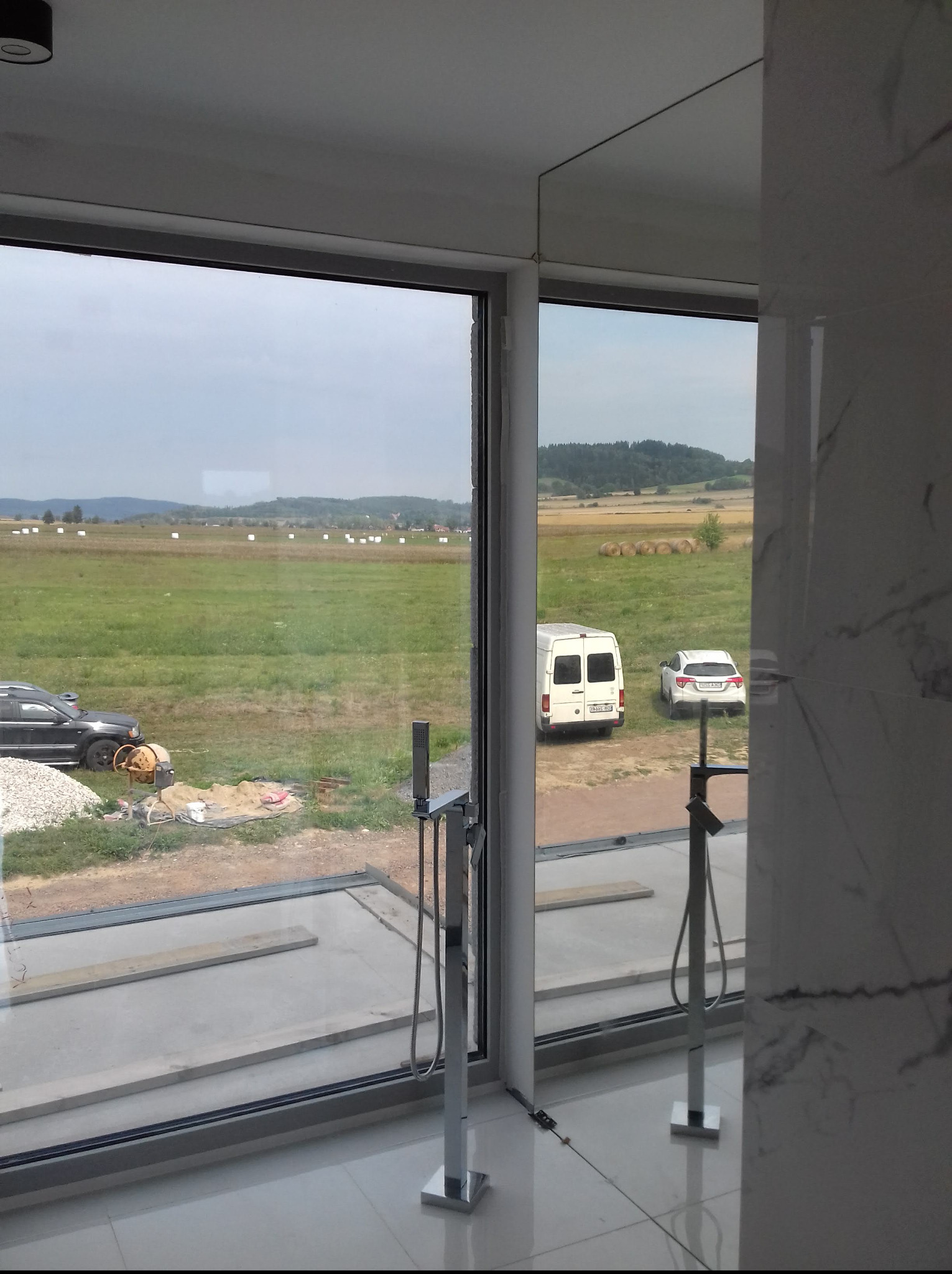 Panoramiczne okno z widokiem na krajobraz, w tle plac budowy z betoniarką. Nowoczesna łazienka z chromowaną armaturą częściowo widoczna.