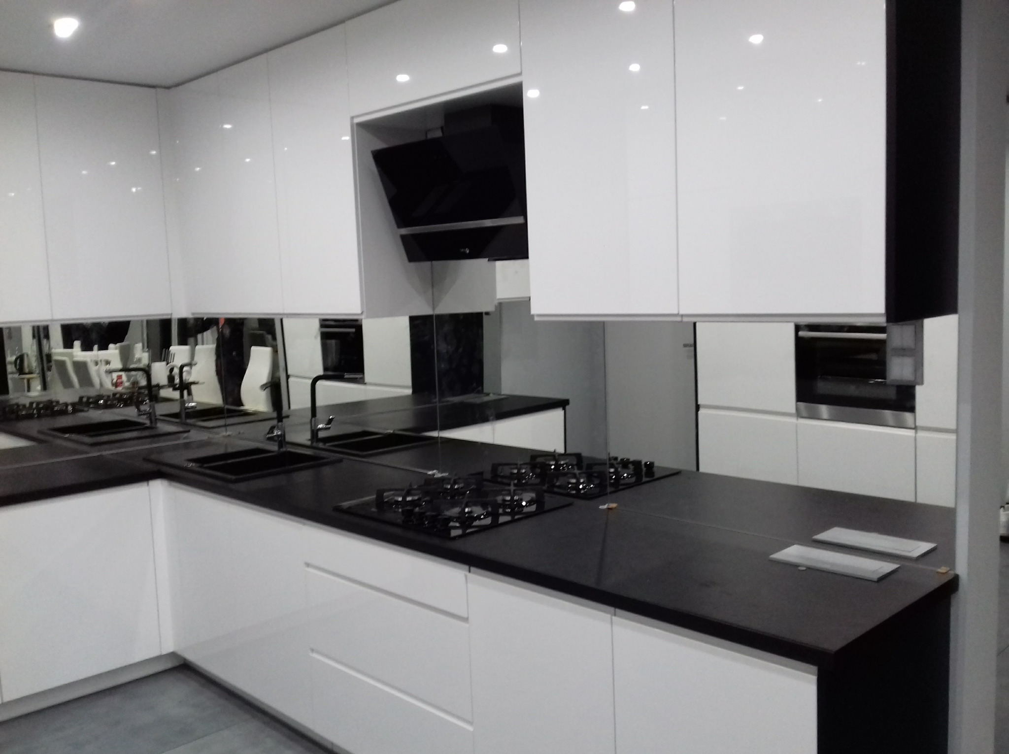 Nowoczesna, biała kuchnia z czarnym blatem i lustrzanymi panelami nad szafkami, odbijającymi przestrzeń. Minimalistyczny design z wbudowanym sprzętem AGD.