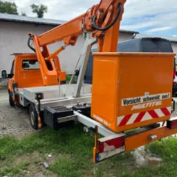 Pomarańczowy podnośnik koszowy Multitel na pojeździe dostawczym, z wysięgnikiem i koszem, zaparkowany na trawiastym terenie. Widoczne tablice ostrzegawcze.