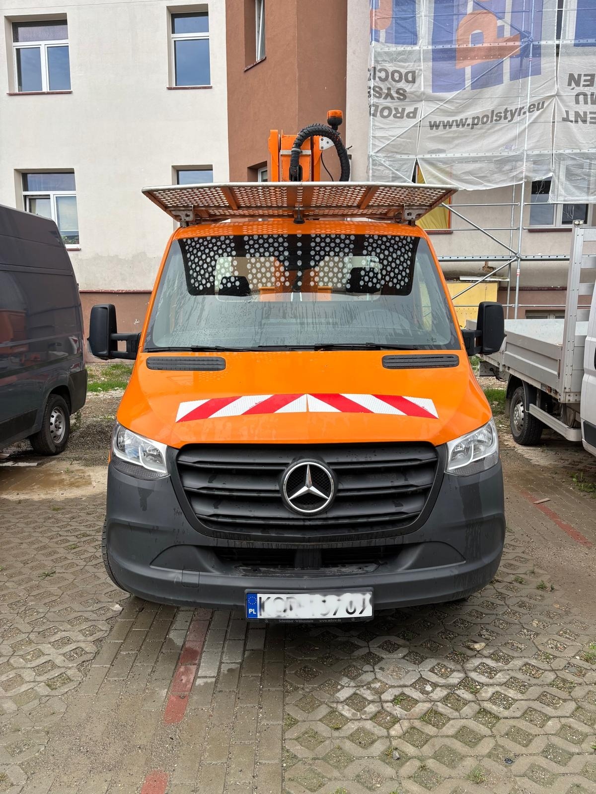Pomarańczowy samochód dostawczy Mercedes z platformą roboczą na dachu, zaparkowany na brukowanej powierzchni przed budynkiem w rusztowaniach. Widok z przodu.