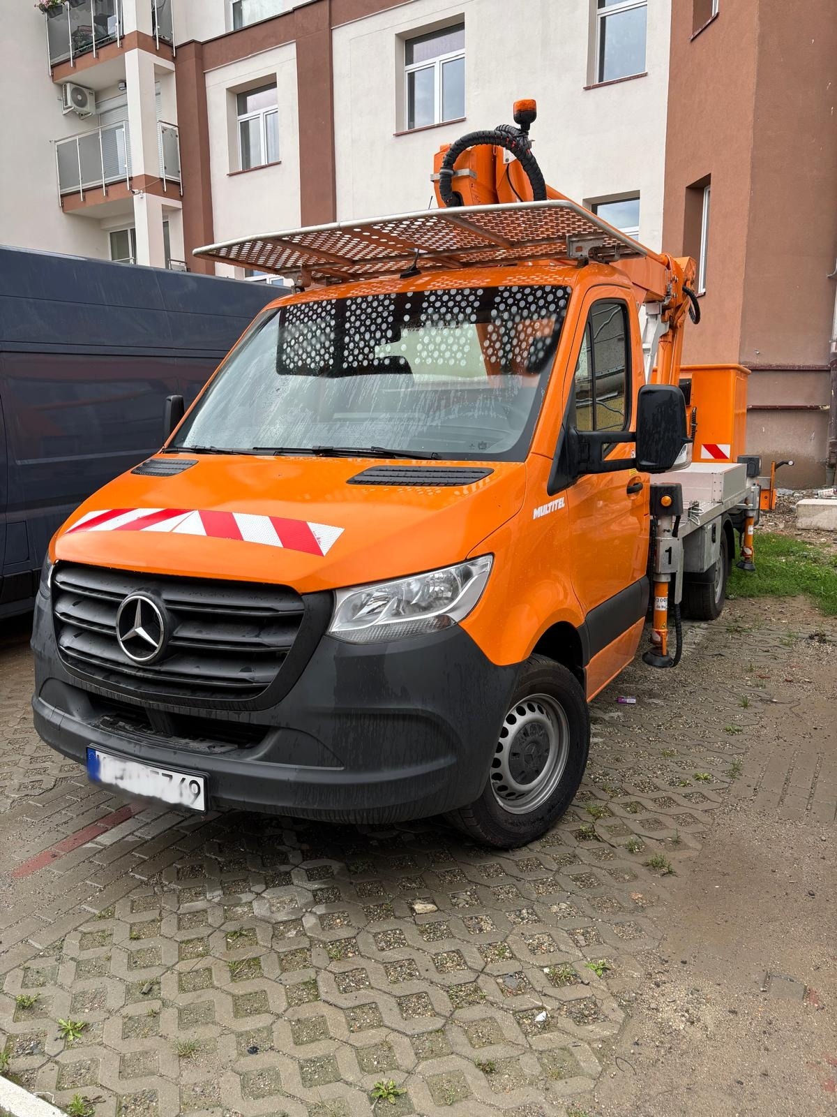 Pomarańczowy samochód z podnośnikiem koszowym marki Mercedes, zaparkowany na brukowanej powierzchni, na tle budynku mieszkalnego. Widoczne detale konstrukcji podnośnika.