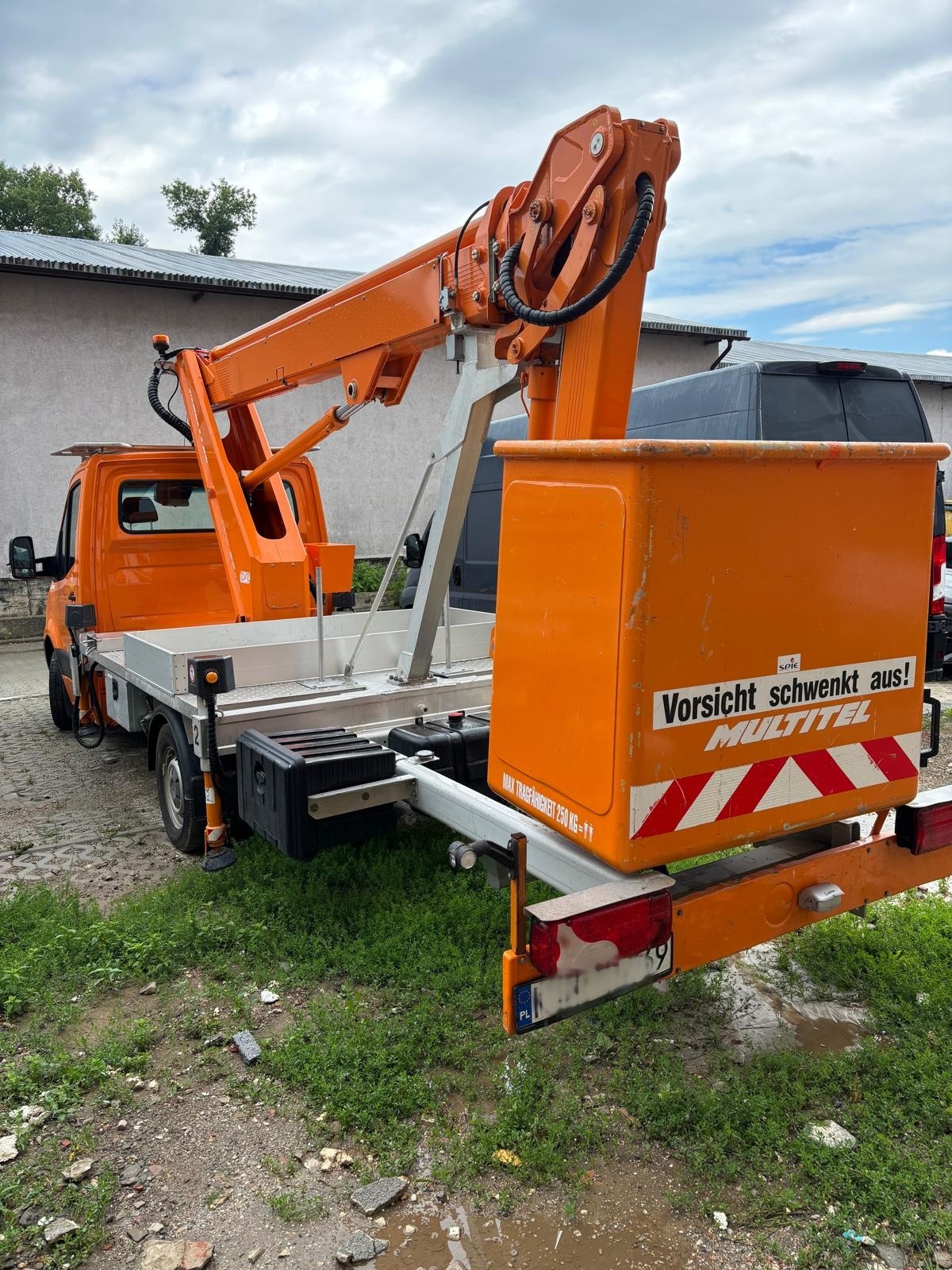 Pomarańczowy podnośnik koszowy Multitel na pojeździe dostawczym, z wysięgnikiem i koszem, zaparkowany na trawiastym terenie. Widoczne tablice ostrzegawcze.