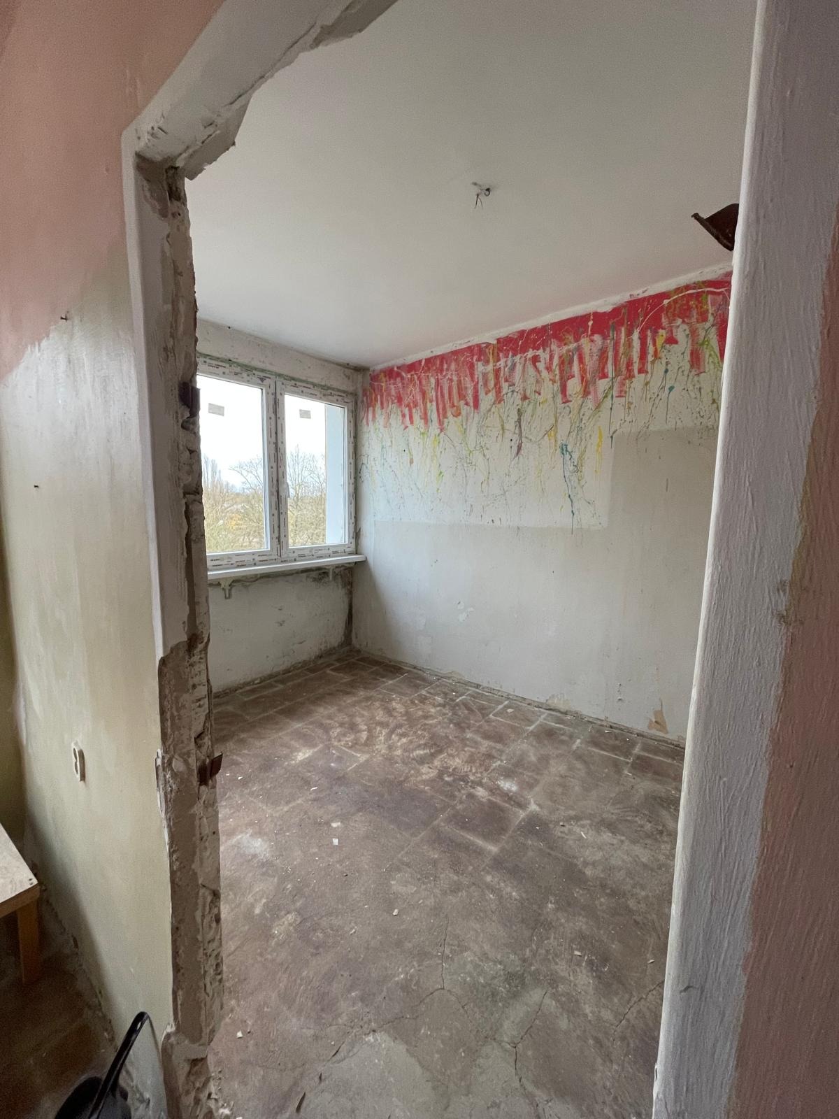 Pokój w trakcie remontu: ściany z resztkami farby, surowa podłoga, widoczne ślady po demontażu. Okno z widokiem na drzewa. Fragment futryny w stanie surowym.