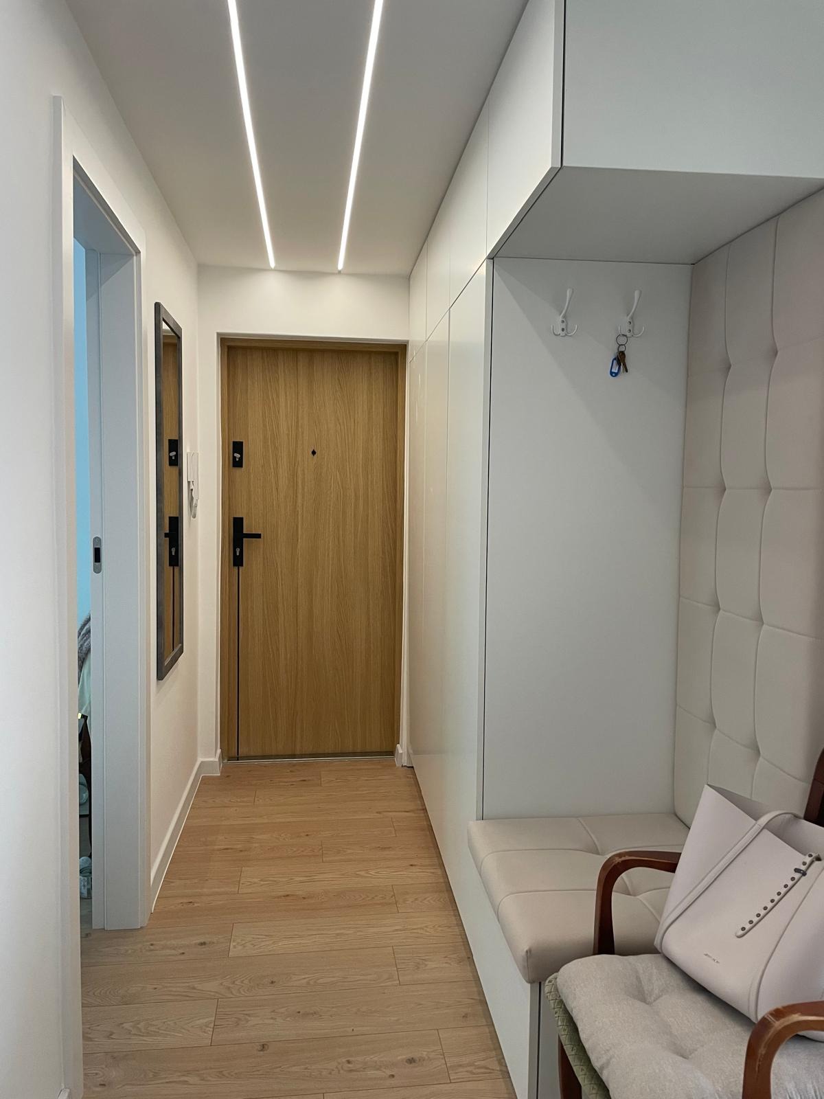 Jasne wnętrze przedpokoju z drewnianą podłogą, białymi szafami w zabudowie i siedziskiem. Dwa pasy ledowe oświetlają przestrzeń. Minimalistyczny design.