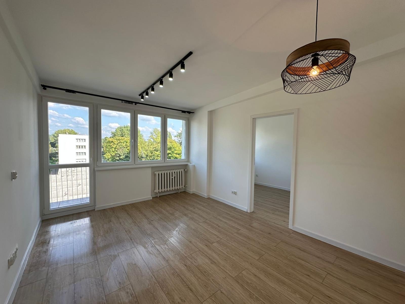 Jasne wnętrze po remoncie: panele podłogowe imitujące drewno, białe ściany, okno z widokiem na zieleń, nowoczesne oświetlenie sufitowe i designerska lampa wisząca.