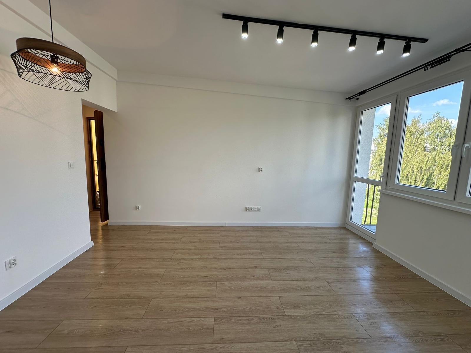 Jasne wnętrze po remoncie: panele podłogowe, białe ściany, okno z widokiem na zieleń, nowoczesne oświetlenie sufitowe i designerska lampa wisząca.