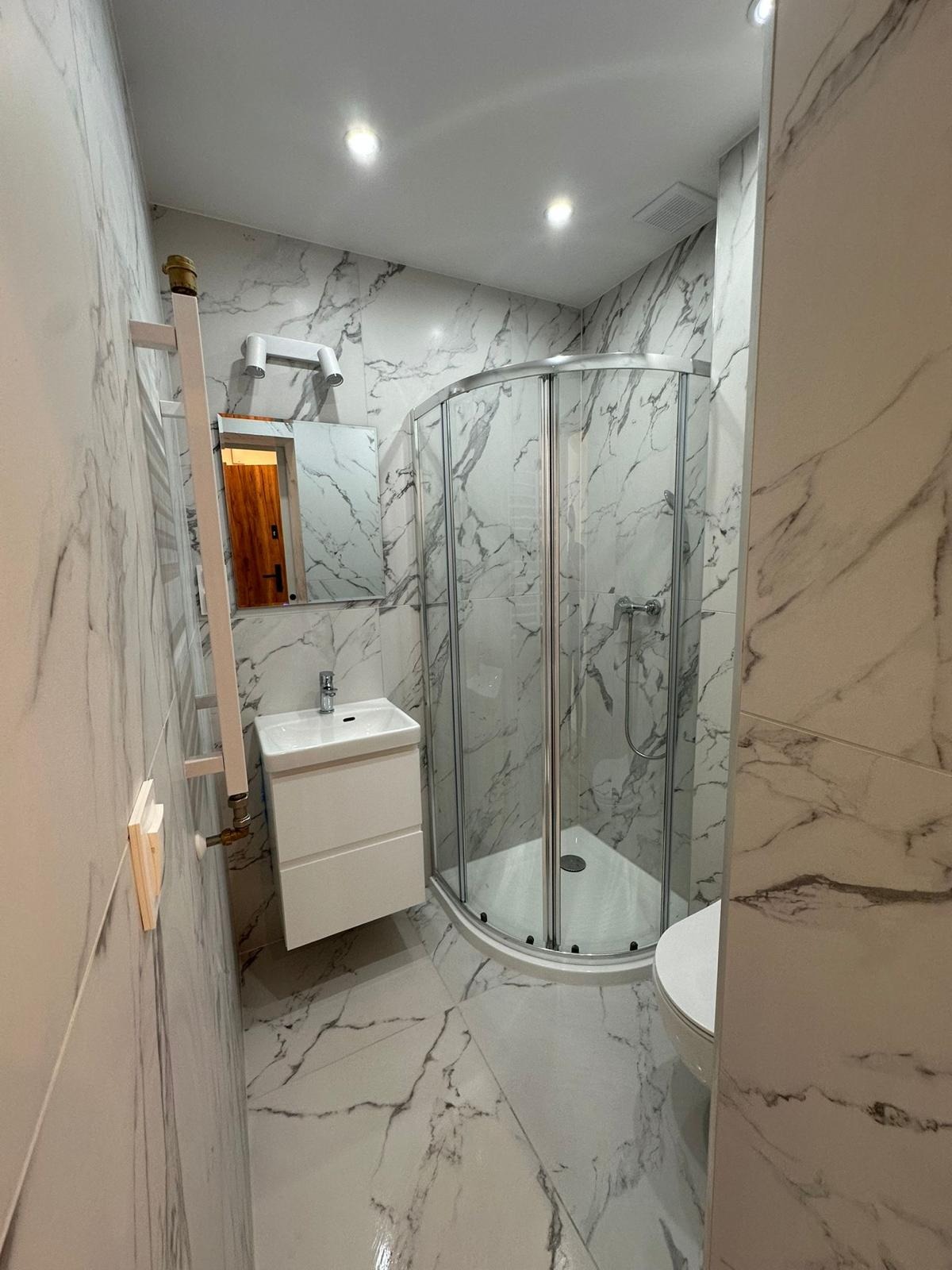 Wykończona łazienka z kabiną prysznicową, umywalką z szafką i lustrem. Białe płytki z szarym marmurkowym wzorem na ścianach i podłodze. Nowoczesne oświetlenie.