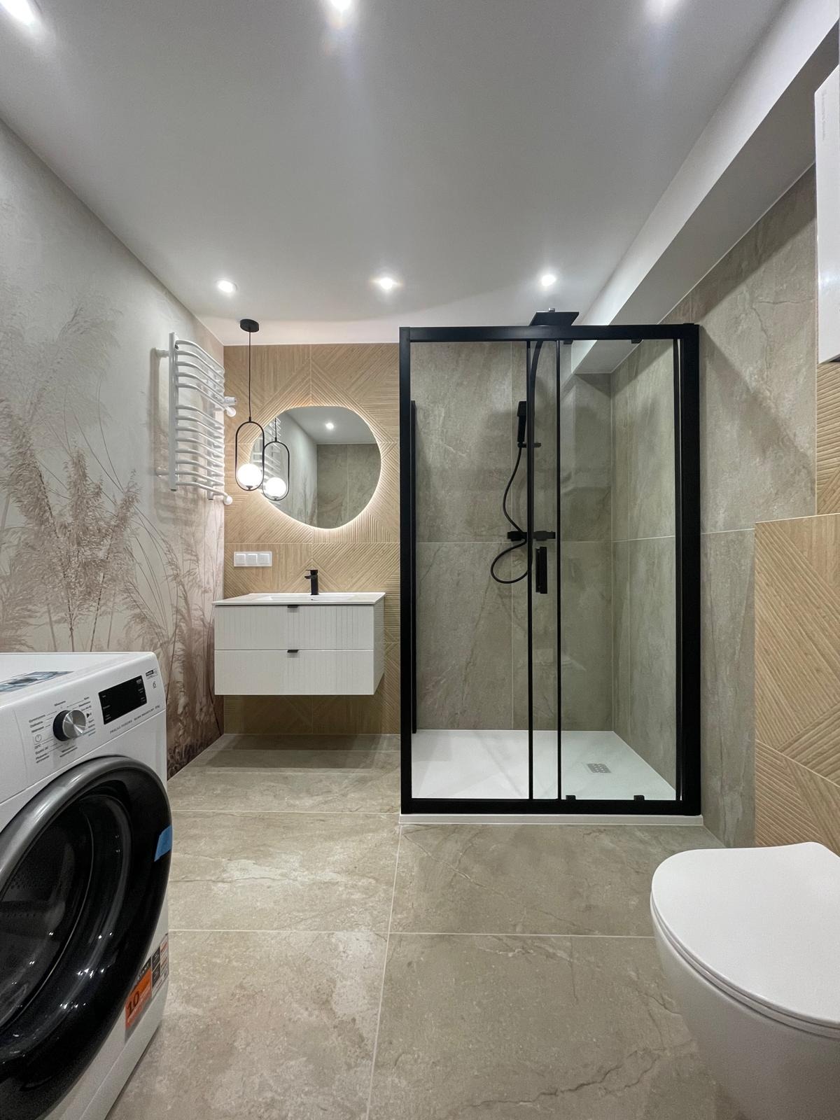 Nowoczesna łazienka z kabiną prysznicową w czarnej ramie, okrągłym lustrem z podświetleniem i pralką. Beżowe płytki i minimalistyczne wykończenie.