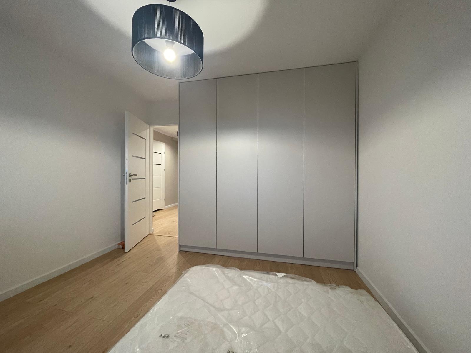 Jasne wnętrze pokoju z parkietem, otwartymi drzwiami i dużą, szarą szafą wnękową. Na podłodze materac zabezpieczony folią. W tle widoczne kolejne drzwi.