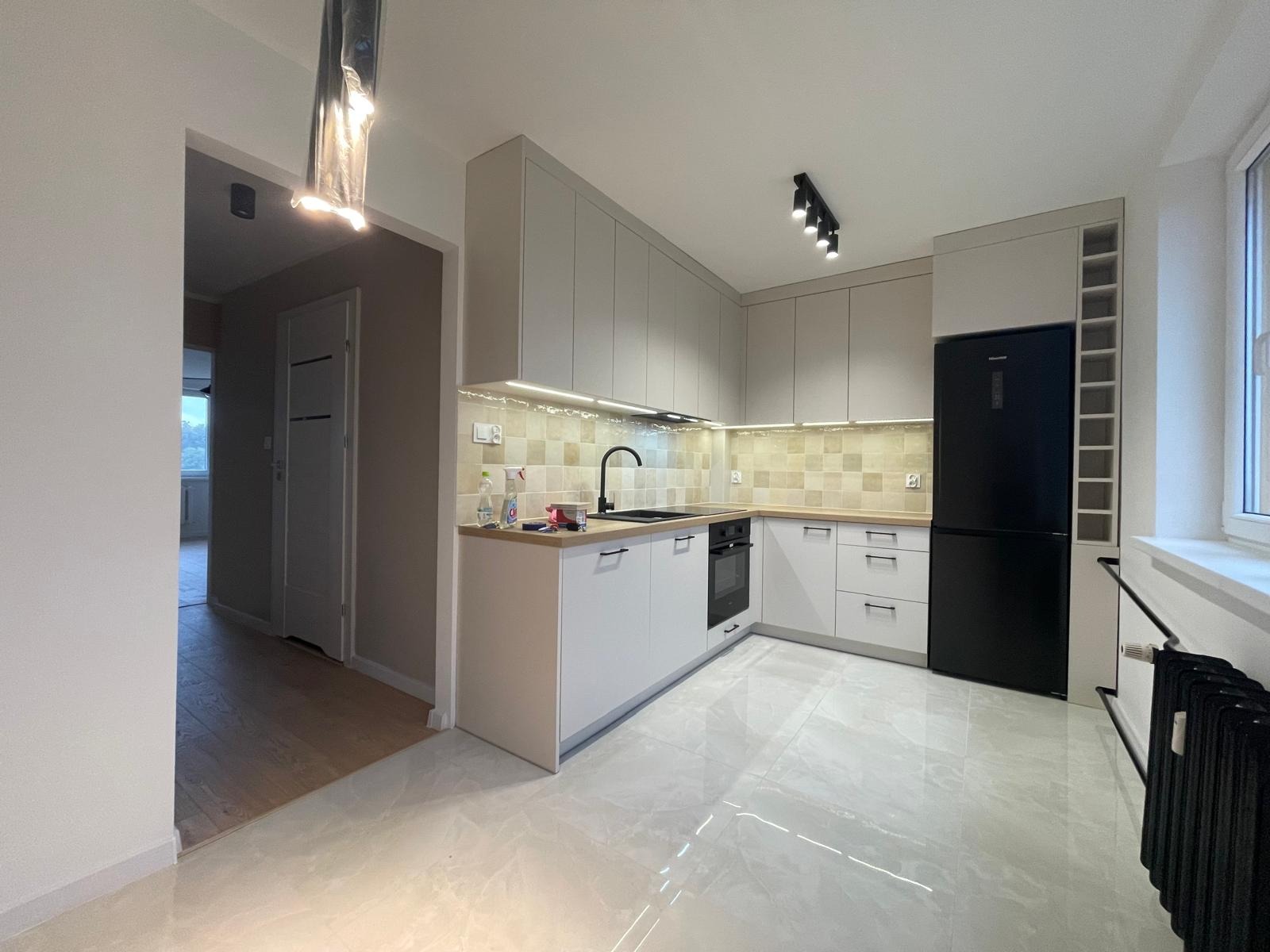 Nowoczesna kuchnia z szarymi szafkami, czarną lodówką i armaturą. Jasne płytki na ścianie i podłodze. Oświetlenie punktowe i listwy LED. Widok na przedpokój z drewnianą podłogą.