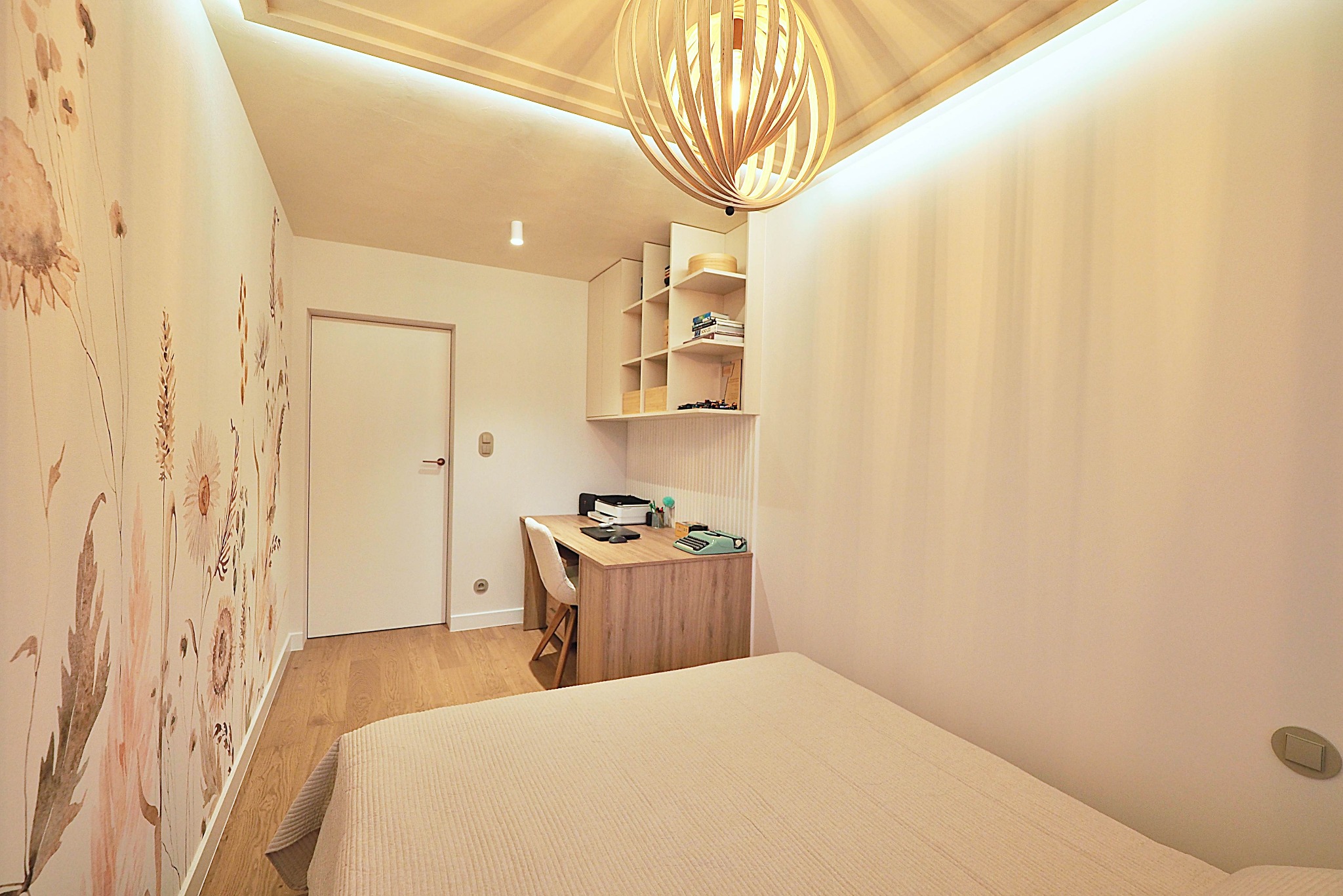 Jasne wnętrze pokoju z łóżkiem, biurkiem i kwiatowym motywem na ścianie. Dekoracyjna lampa sufitowa i subtelne oświetlenie LED dodają przytulności.