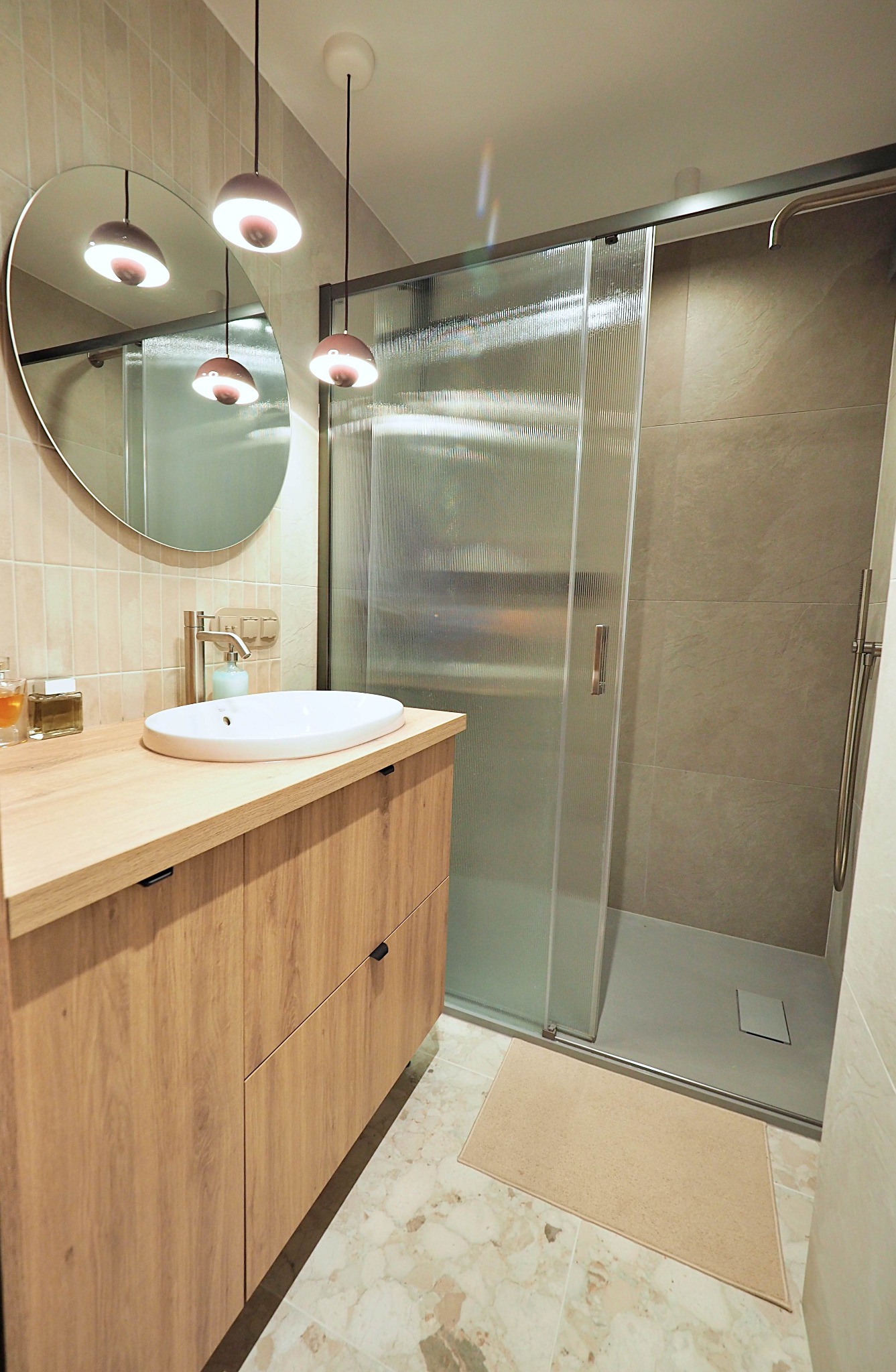 Nowoczesna łazienka z drewnianą szafką pod umywalką, okrągłym lustrem i kabiną prysznicową z ryflowanymi szybami. Uwagę przykuwają designerskie lampy wiszące.