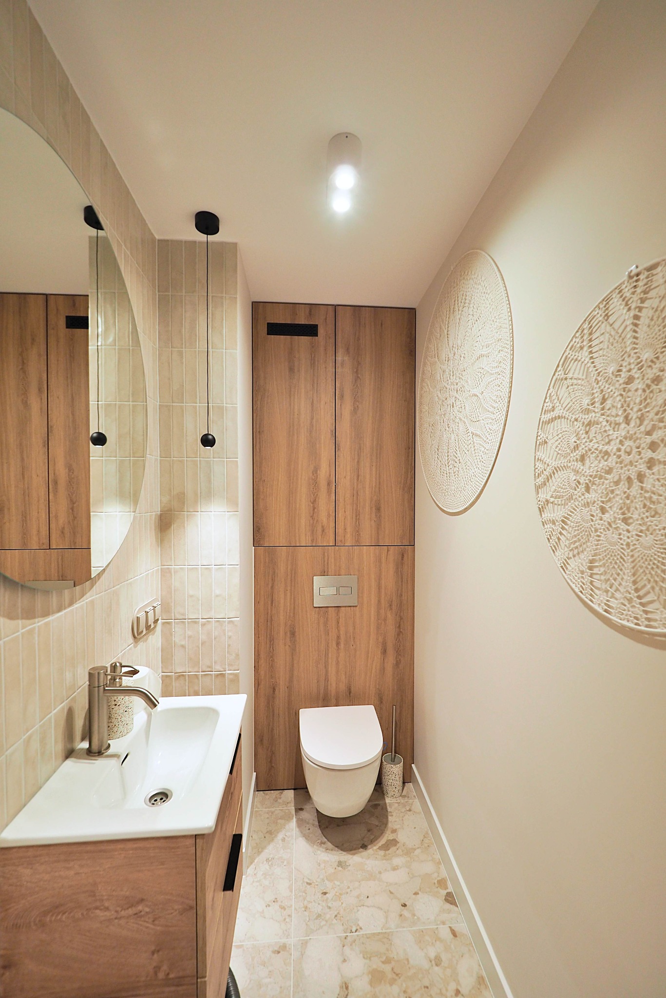 Nowoczesna aranżacja małej łazienki w Koszalinie. Umywalka z szafką z drewnianym dekorem, okrągłe lustro, beżowe płytki i dekoracje ścienne.