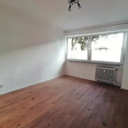 Jasne wnętrze po remoncie: drewniana podłoga, białe ściany, grzejnik pod oknem z widokiem na zieleń. Minimalistyczna lampa sufitowa.