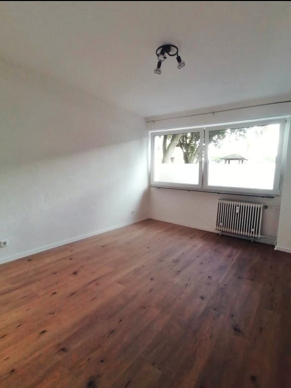 Jasne wnętrze po remoncie: drewniana podłoga, białe ściany, grzejnik pod oknem z widokiem na zieleń. Minimalistyczna lampa sufitowa.