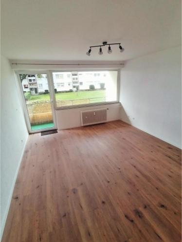 Jasne wnętrze pokoju z drewnianą podłogą, grzejnikiem pod oknem i nowoczesnym oświetleniem sufitowym z czterema żarówkami. Widok na zieleń za oknem.