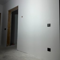 TWÓJ PARTNER GROUP SPÓŁKA Z OGRANICZONĄ ODPOWIEDZIALNOŚCIĄ - Świeżo pomalowane, białe ściany z czarnymi włącznikami, surowa ościeżnica i betonowa podłoga w nowym wnętrzu. Widoczny otwór po gniazdku.