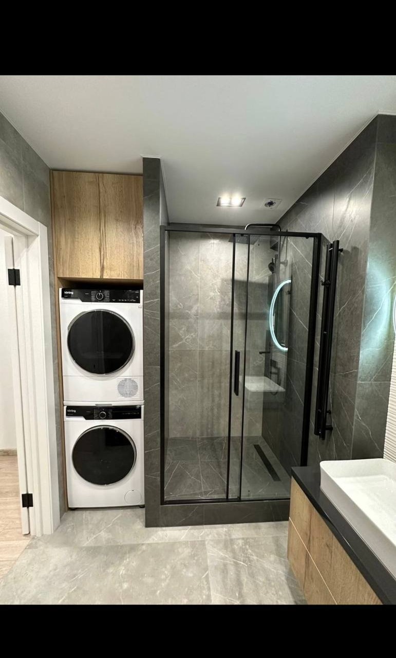 Nowoczesna łazienka z kabiną prysznicową typu walk-in, pralką i suszarką w zabudowie oraz umywalką z szafką, utrzymana w szarościach i drewnie.