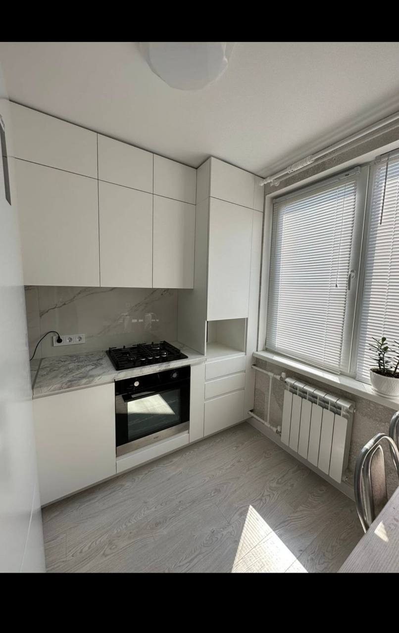 Jasna kuchnia z białymi szafkami, marmurowym blatem i drewnianą podłogą. Okno z żaluzjami wpuszcza światło na grzejnik. Minimalistyczny design.