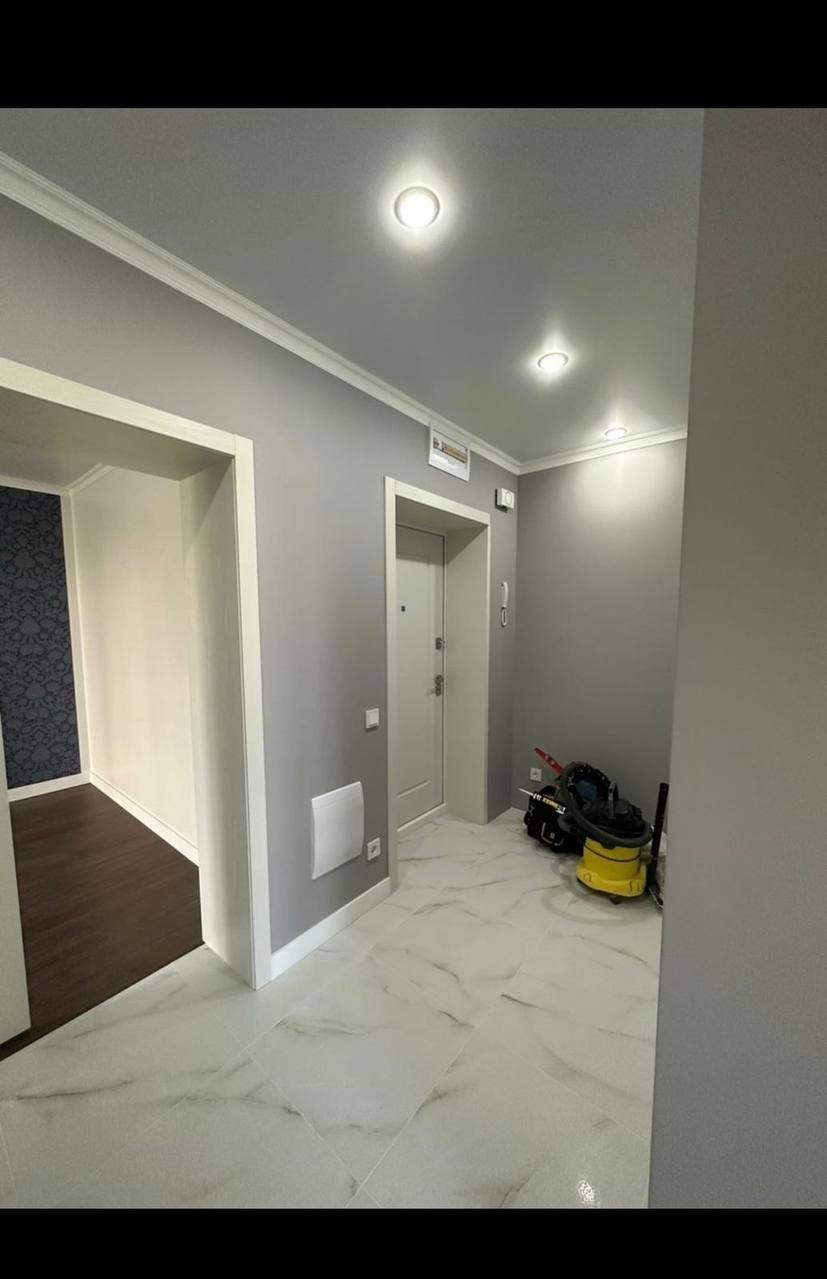 Korytarz po odnowieniu: szare ściany z białą sztukaterią, marmurowe płytki podłogowe, drzwi wejściowe i widok na pokój z ciemną podłogą. Widoczny odkurzacz.