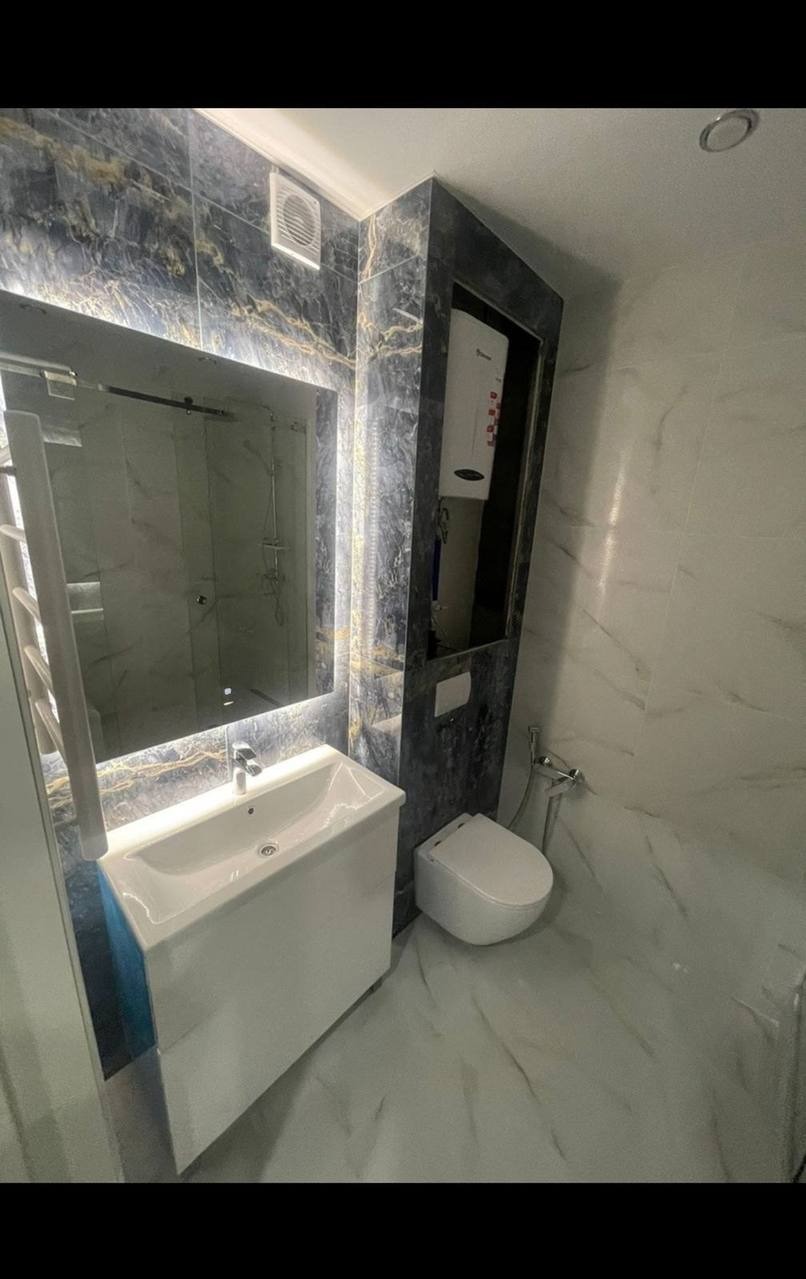 Elegancka, wykończona łazienka w bieli i granacie z podświetlanym lustrem, umywalką z szafką i toaletą. Widoczny bojler w zabudowie. Marmurowe płytki na podłodze i ścianach.