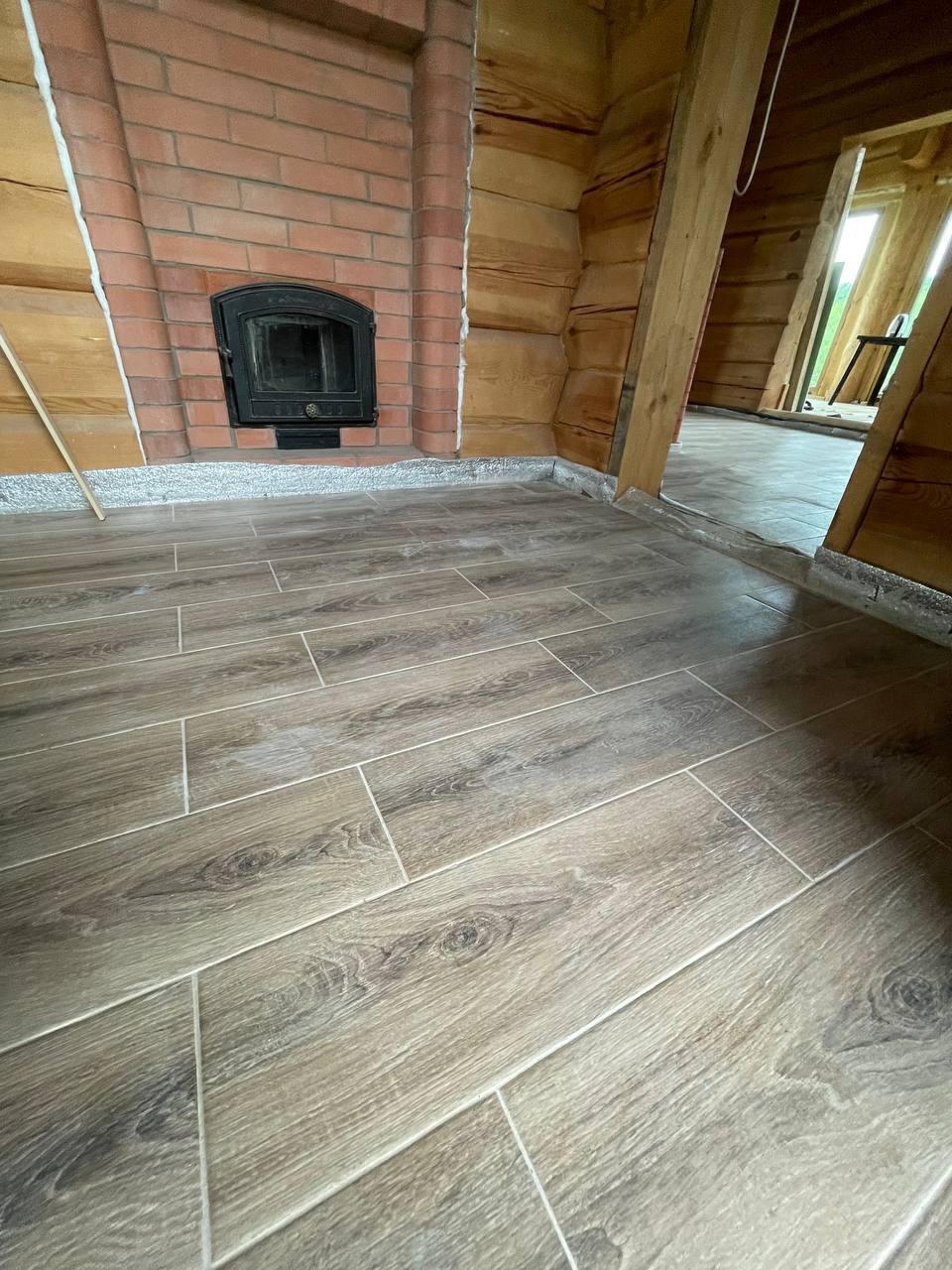 Ukończona podłoga z płytek imitujących drewno w rustykalnym wnętrzu z kominkiem. Widoczny fragment drewnianej ściany i przejście do innego pomieszczenia.