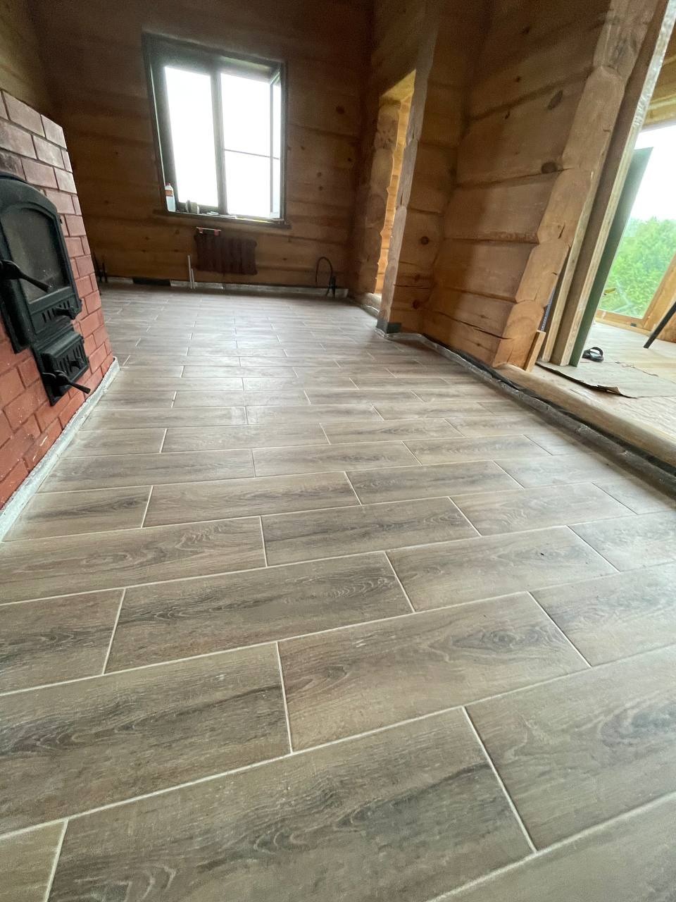 Ukończona podłoga z płytek imitujących drewno w drewnianym domu z kominkiem, widok na okno i drzwi. Jasne fugi. Podłoga ułożona równolegle do ściany.