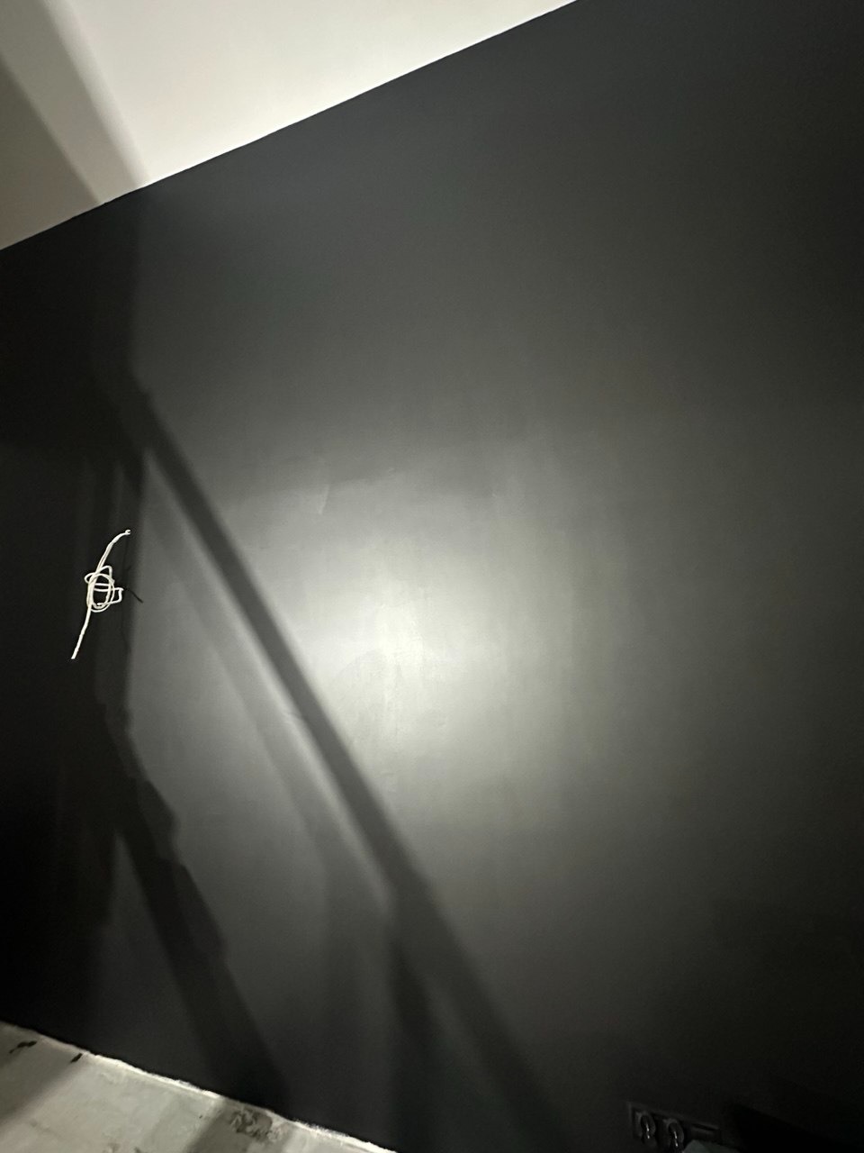 Ściana pomalowana na ciemny kolor z wystającymi przewodami elektrycznymi. Widoczne niedoskonałości malowania, oświetlenie boczne.