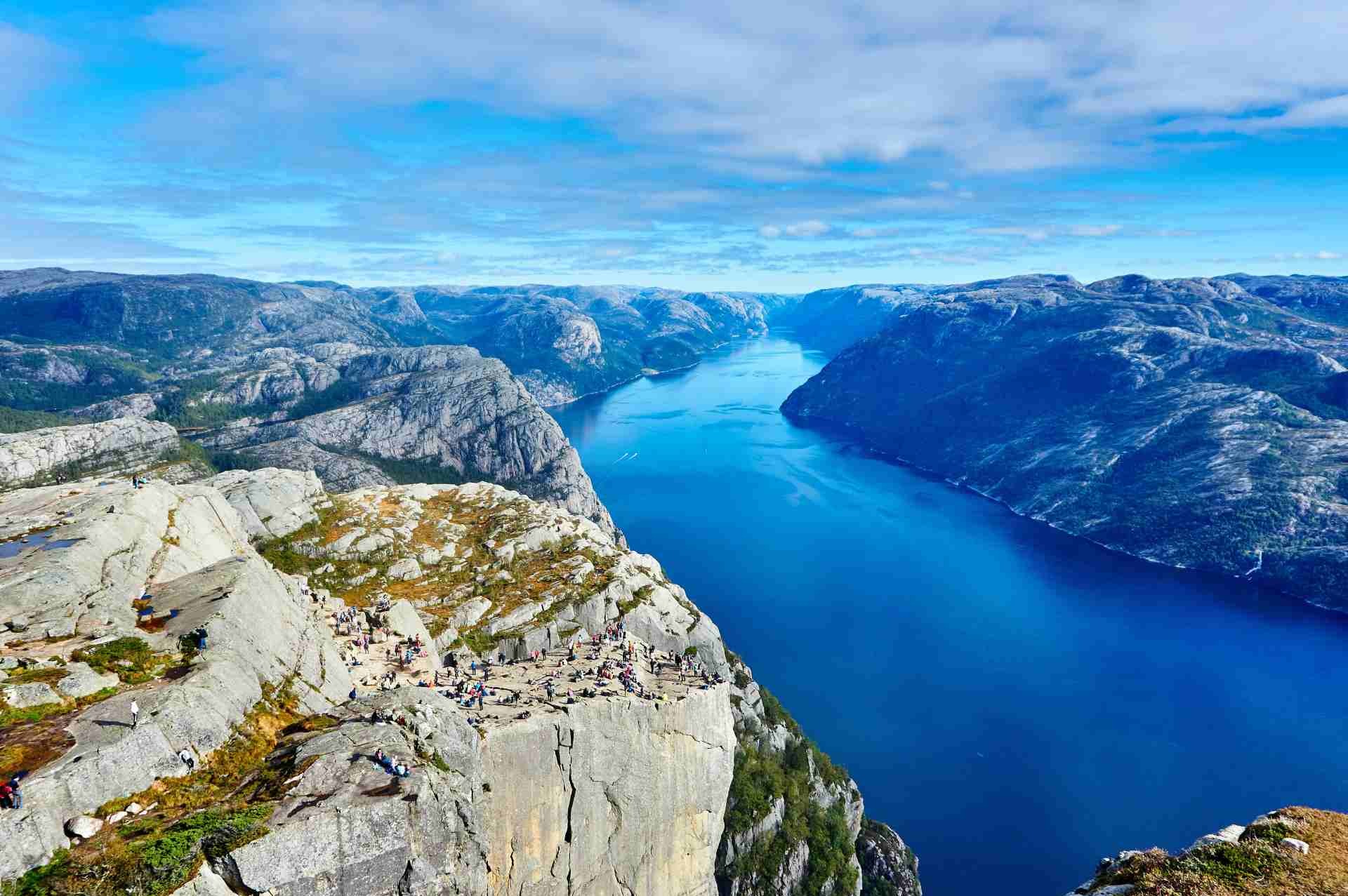 Widok z Preikestolen: klif z grupą turystów, głęboki fiord i górzysty krajobraz w Norwegii pod błękitnym niebem. Panorama z góry.