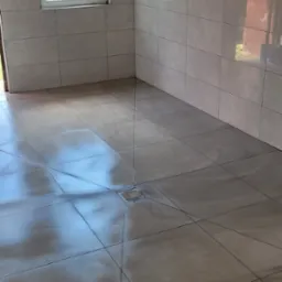 Wykończona łazienka z beżowymi, błyszczącymi płytkami na ścianach i podłodze. Widoczny odpływ liniowy w podłodze i okno z widokiem na zewnątrz.