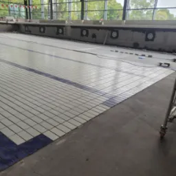 Układanie płytek na dnie basenu z niebieskimi pasami, widok na duże okna i częściowo widoczny rusztowanie. Prace wykończeniowe w toku.