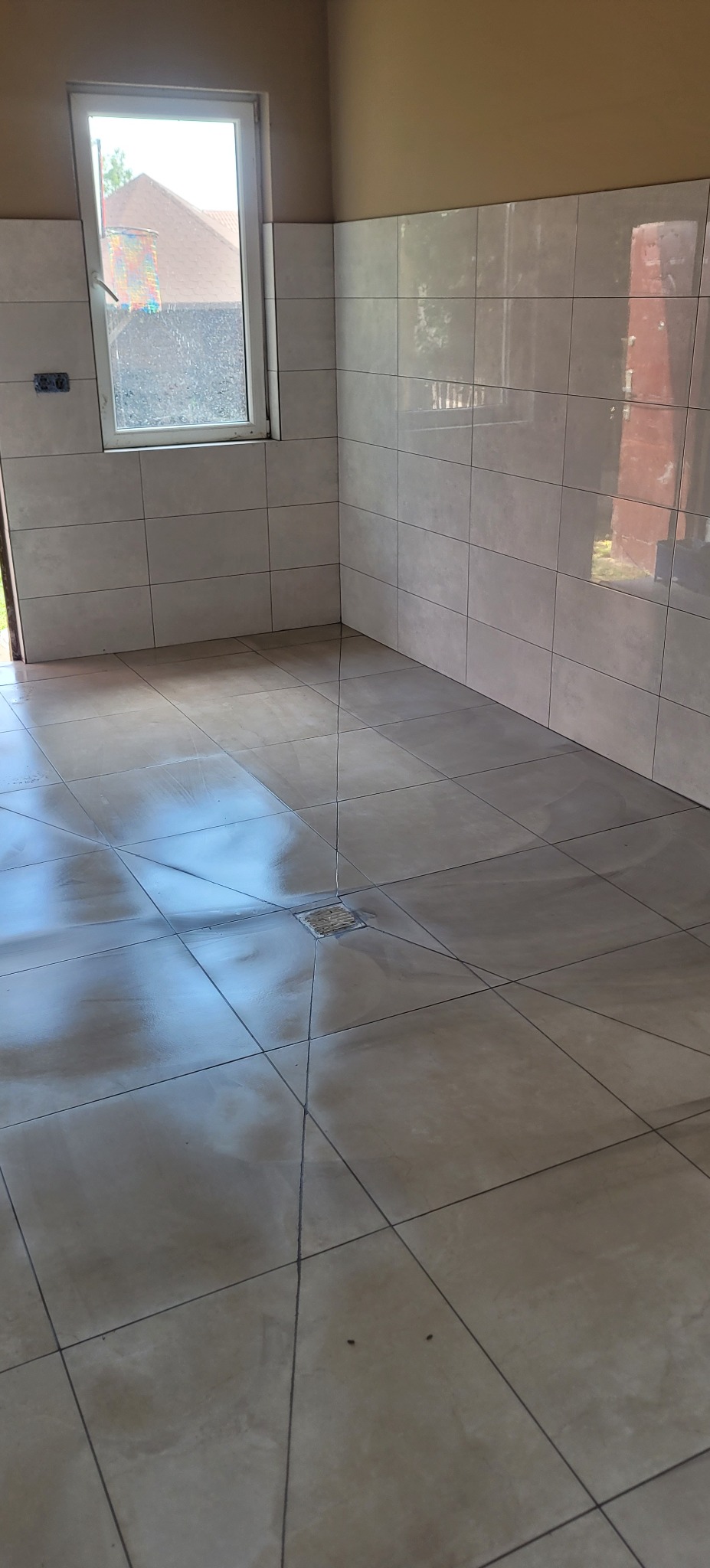 Wykończona łazienka z beżowymi, błyszczącymi płytkami na ścianach i podłodze. Widoczny odpływ liniowy w podłodze i okno z widokiem na zewnątrz.