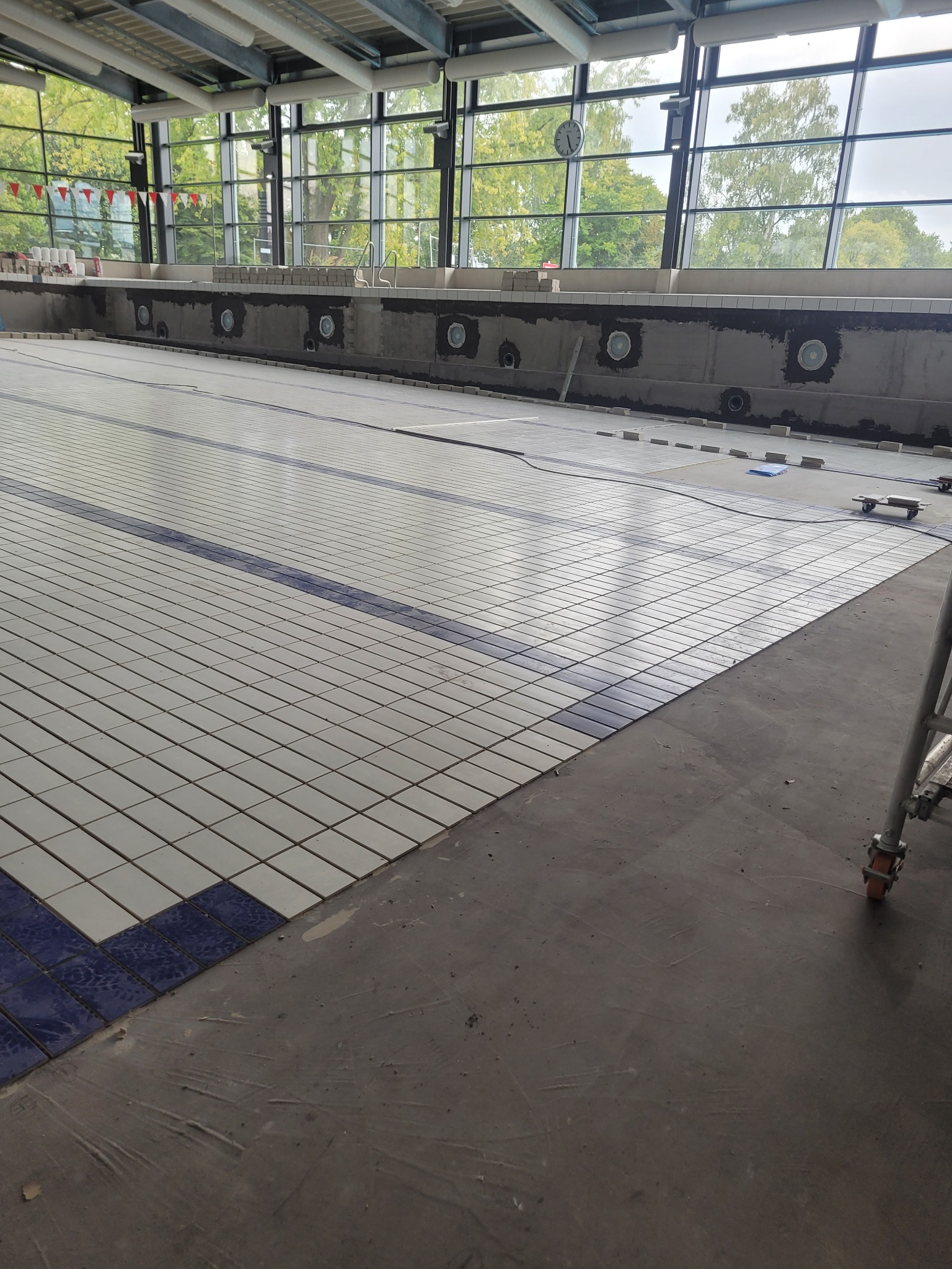 Układanie płytek na dnie basenu z niebieskimi pasami, widok na duże okna i częściowo widoczny rusztowanie. Prace wykończeniowe w toku.